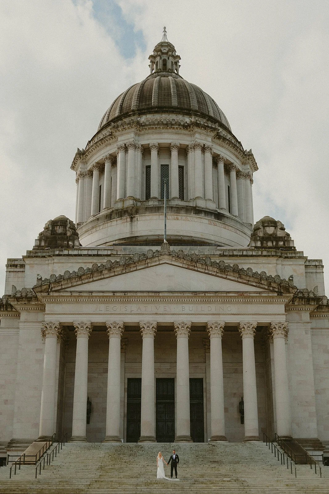 A wedding couple standing on the steps in front of a large, neoclassical building with columns and a domed roof.