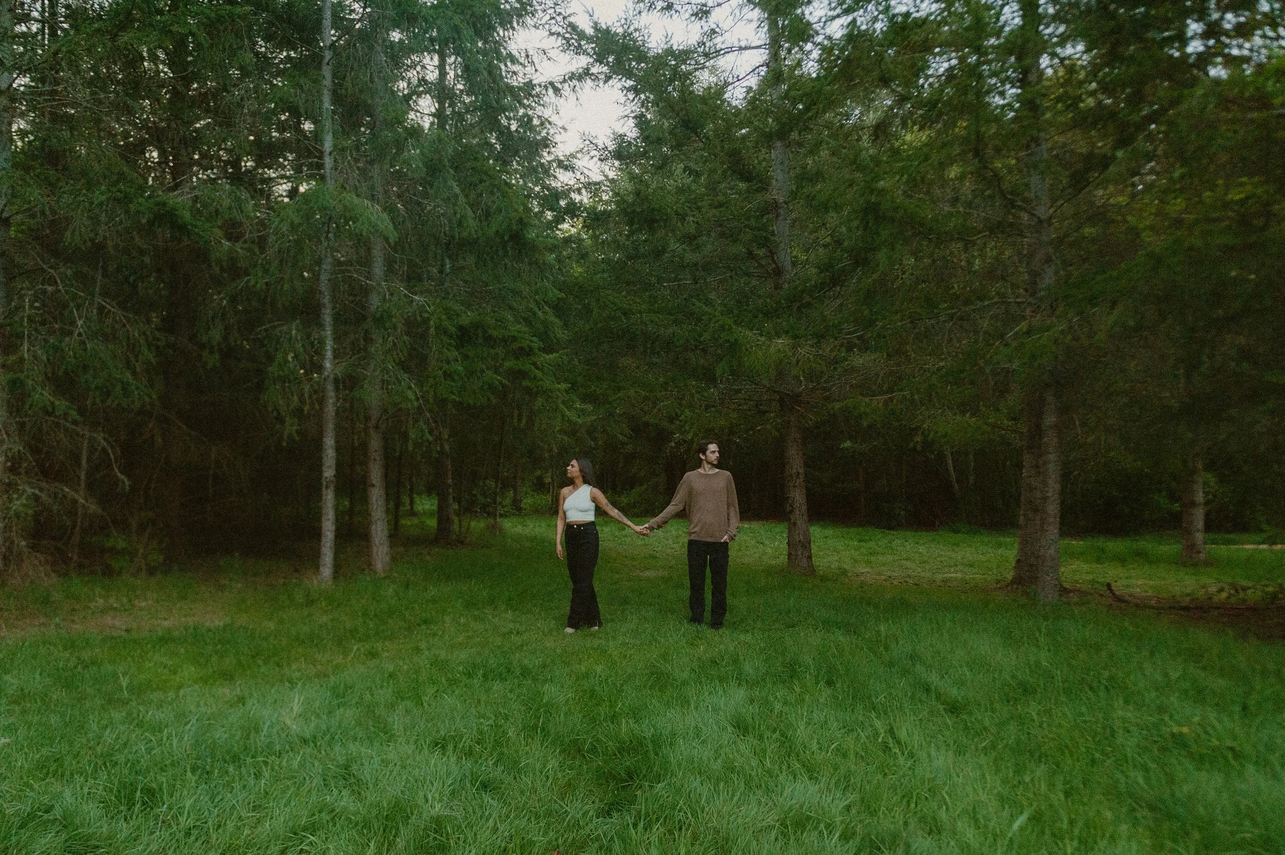 A couple holding hands, walking through a grassy forest clearing surrounded by tall trees.