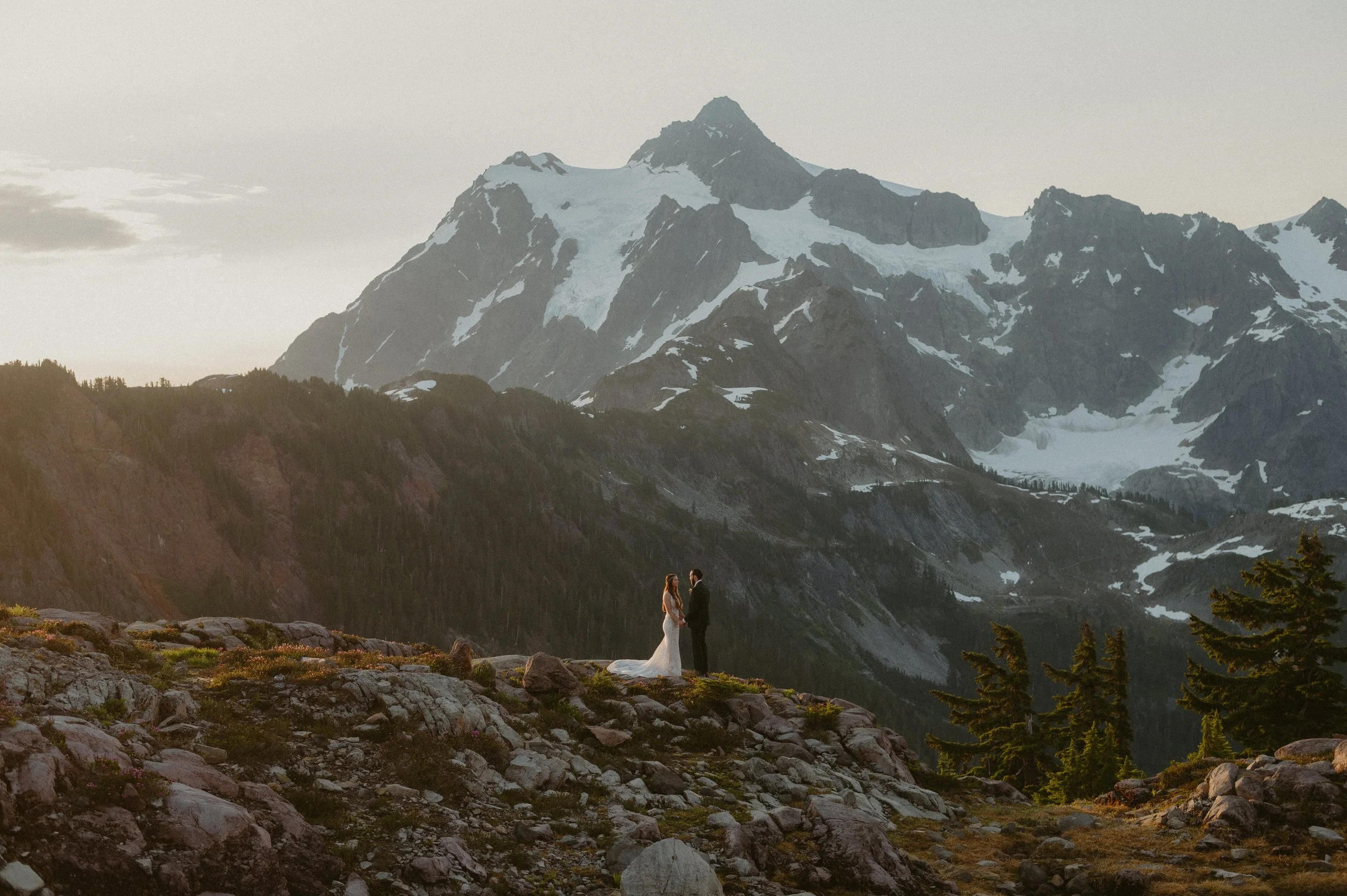 A couple in wedding attire standing on rocky terrain with snow-capped mountains in the background during sunset or sunrise.