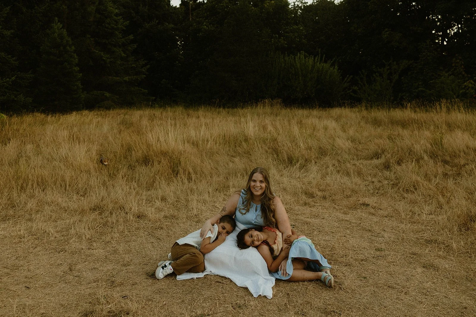 A woman and two children sitting on a white blanket in a field of dry grass, with trees in the background. The woman has long hair and is smiling, while the children are resting against her, with one leaning on her lap and the other lying on her side