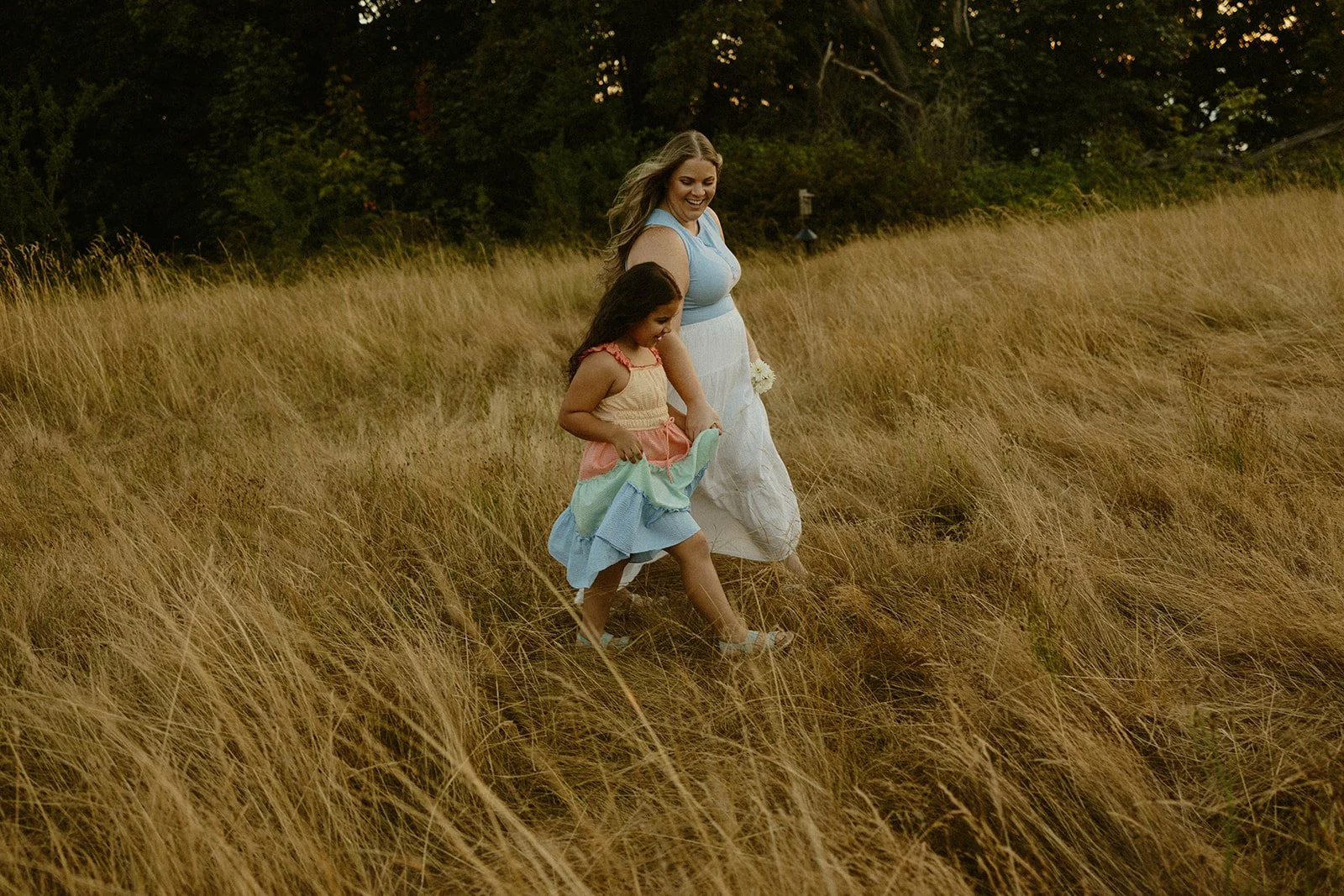 A woman and a young girl walking through a field of tall, golden grass in the evening, with trees in the background.