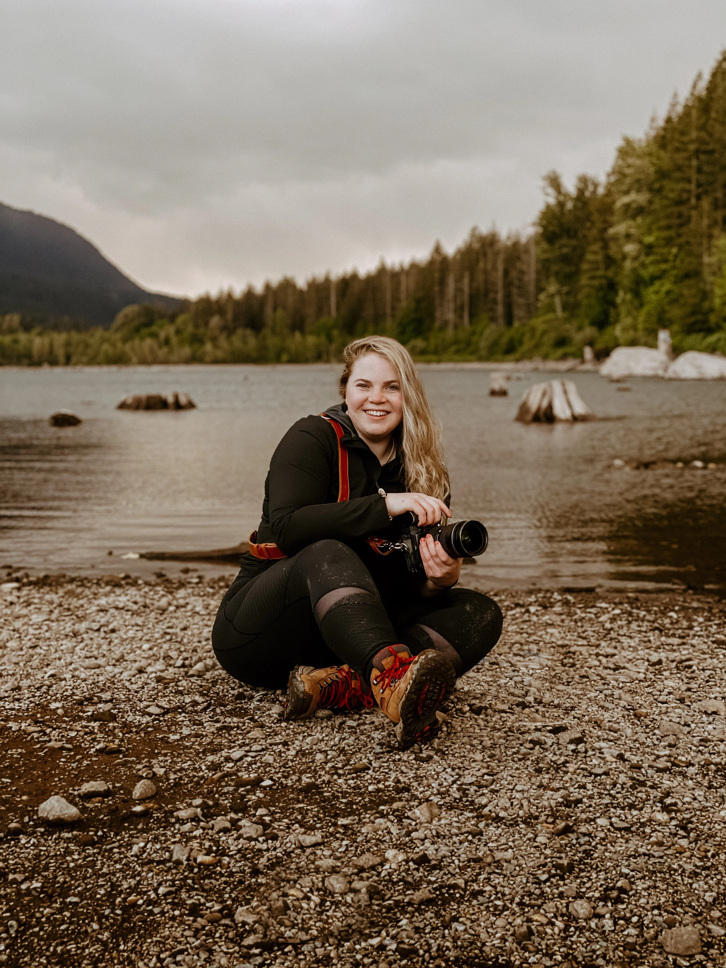 A woman sitting on a rocky shoreline, holding a camera, with a lake, trees, and mountains in the background under an overcast sky.
