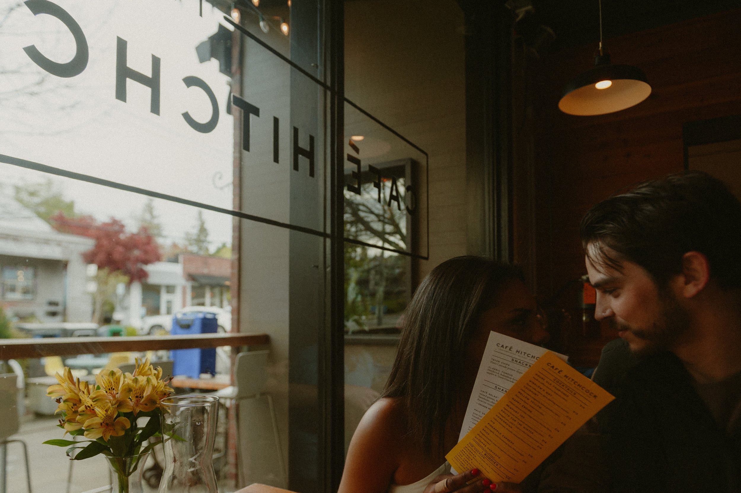 A couple sitting inside a cafe, looking at a menu together near a window with a view of trees and houses outside.