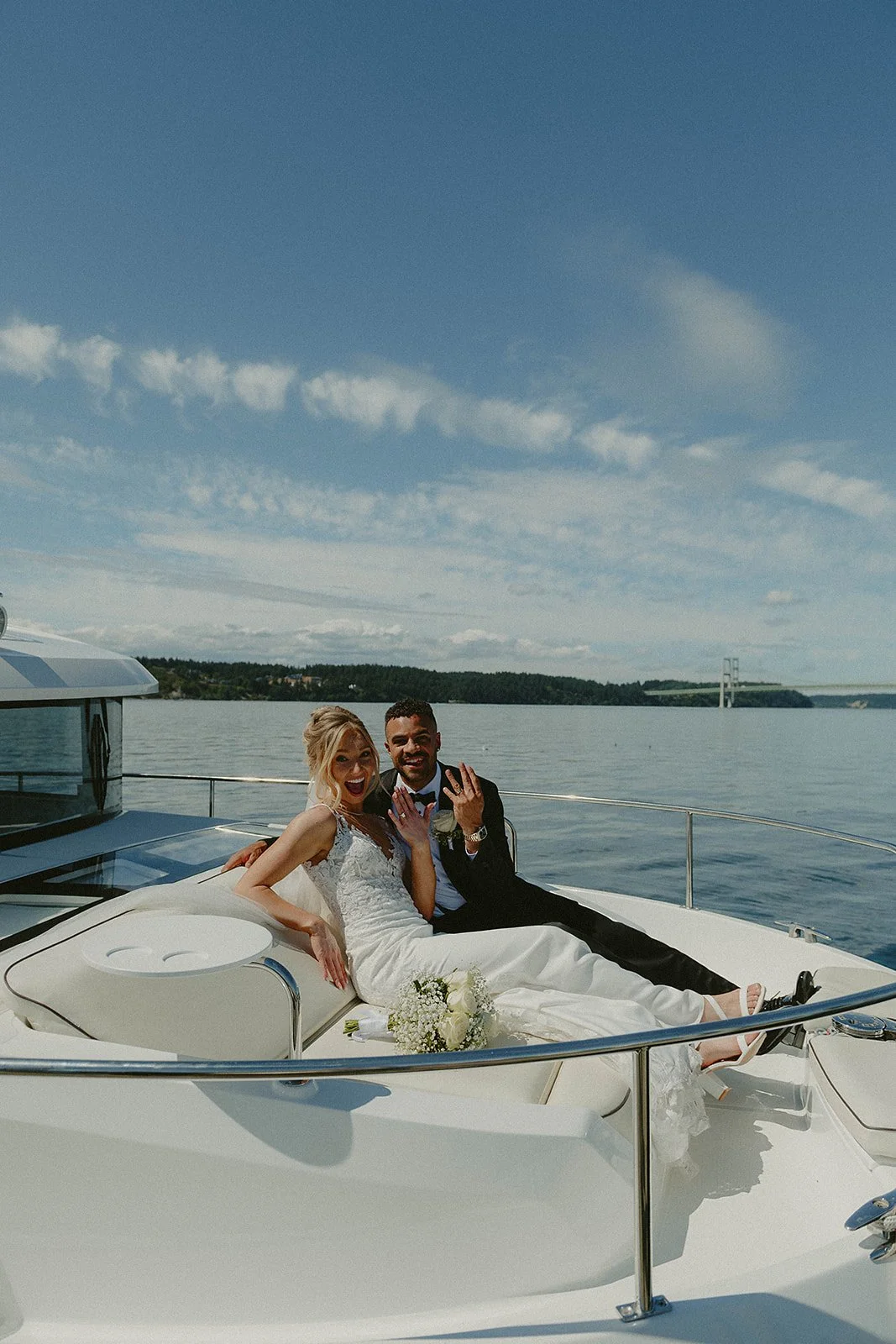 A newlywed couple in wedding attire celebrating on a boat with a scenic water view and bridge in the background.