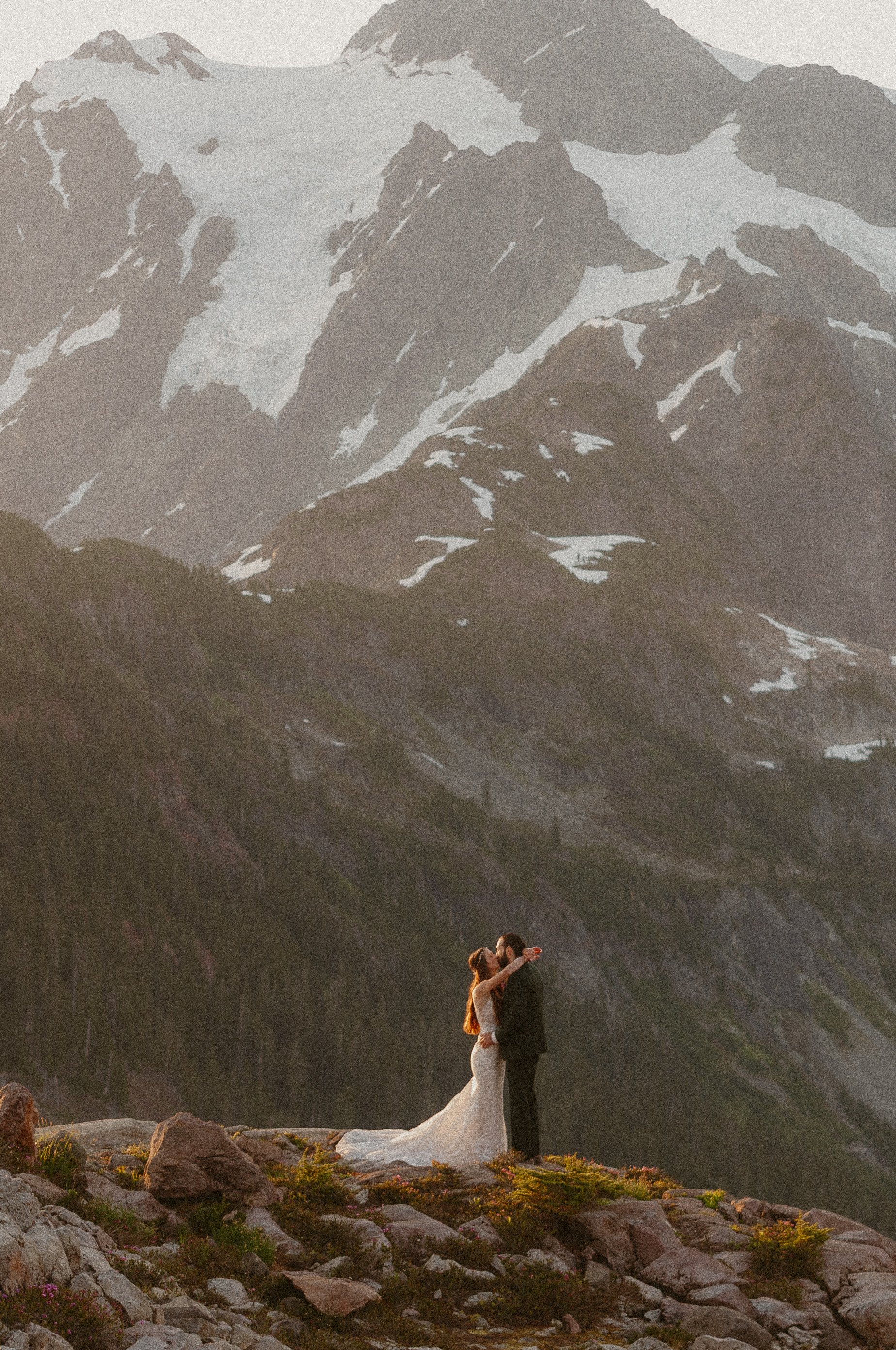 A bride and groom embrace and kiss in a mountainous landscape with snow-capped peaks in the background, rocky terrain in the foreground.