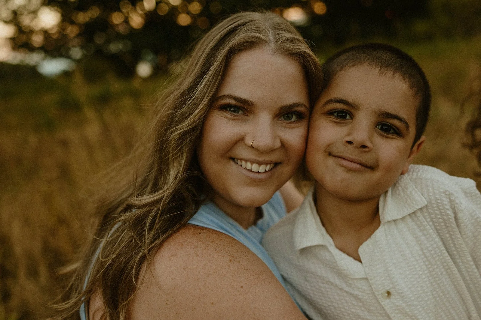 A woman and a young boy smiling and leaning their heads together outdoors during sunset.