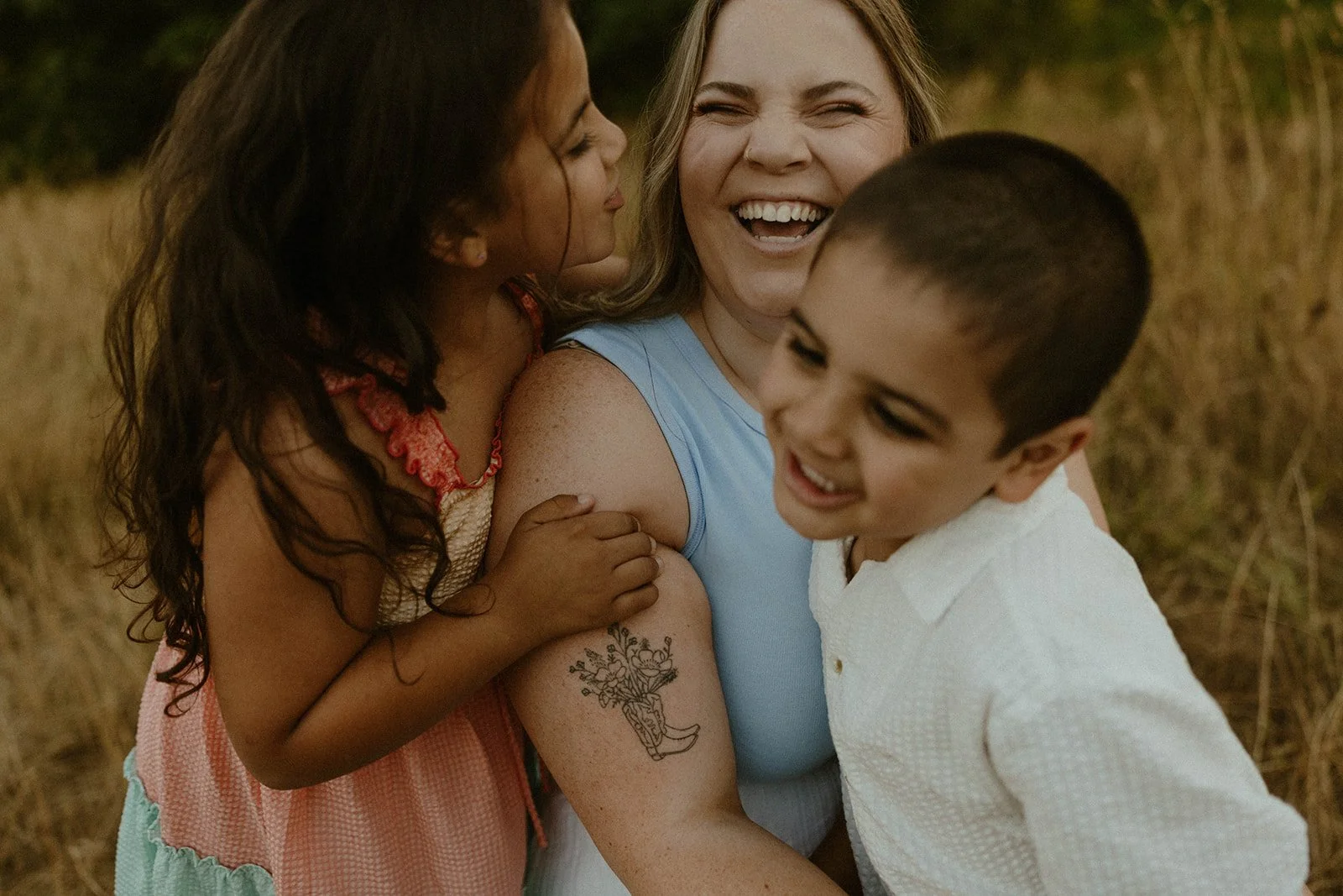 Three people, a woman with blonde hair, a girl with dark curly hair, and a boy with short dark hair, are outside in a grassy field, smiling and hugging each other, enjoying a happy moment together.