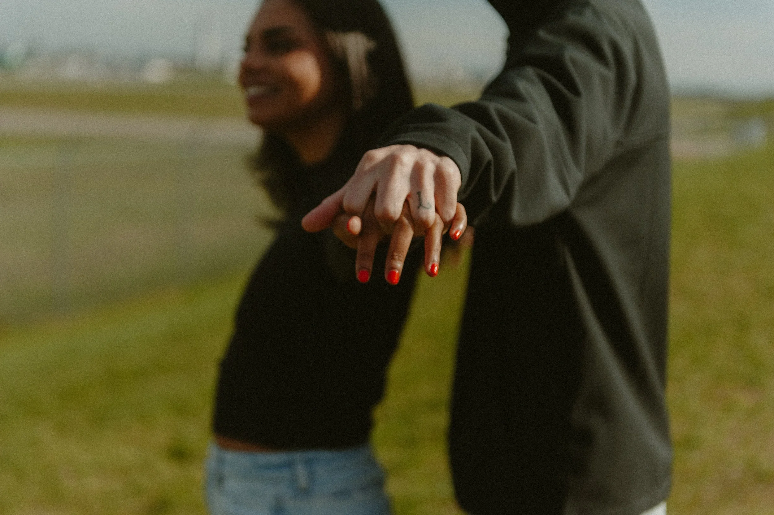 A woman with long black hair and red nails is holding hands with a person outside in a green field. The woman's face is partly visible, and both are smiling. The person is wearing a dark long-sleeve shirt, and the woman is wearing a black top.