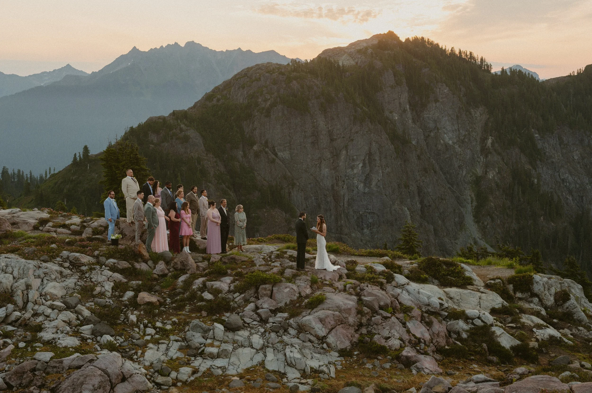 A wedding ceremony taking place outdoors on rocky terrain with a mountain backdrop at sunset. The bride and groom stand facing each other, exchanging vows, while guests stand on a slight incline behind them, dressed in formal attire.