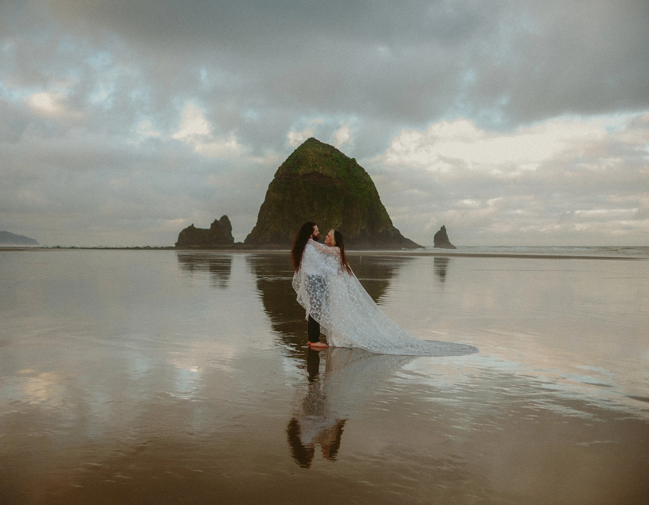 Two women in wedding dresses standing on a beach, facing each other, with their reflection visible in the wet sand, in front of a large rock formation and smaller rocks in the background, under a cloudy sky.