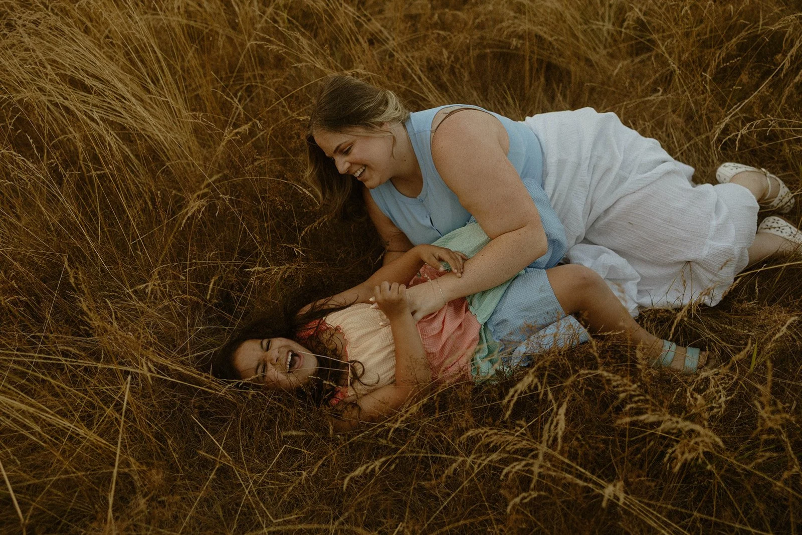 A woman and a young girl are playing and laughing on a grassy field. The woman is on her side, leaning over the girl, who is lying on her back among the tall grass. They are smiling and holding hands, enjoying each other's company.