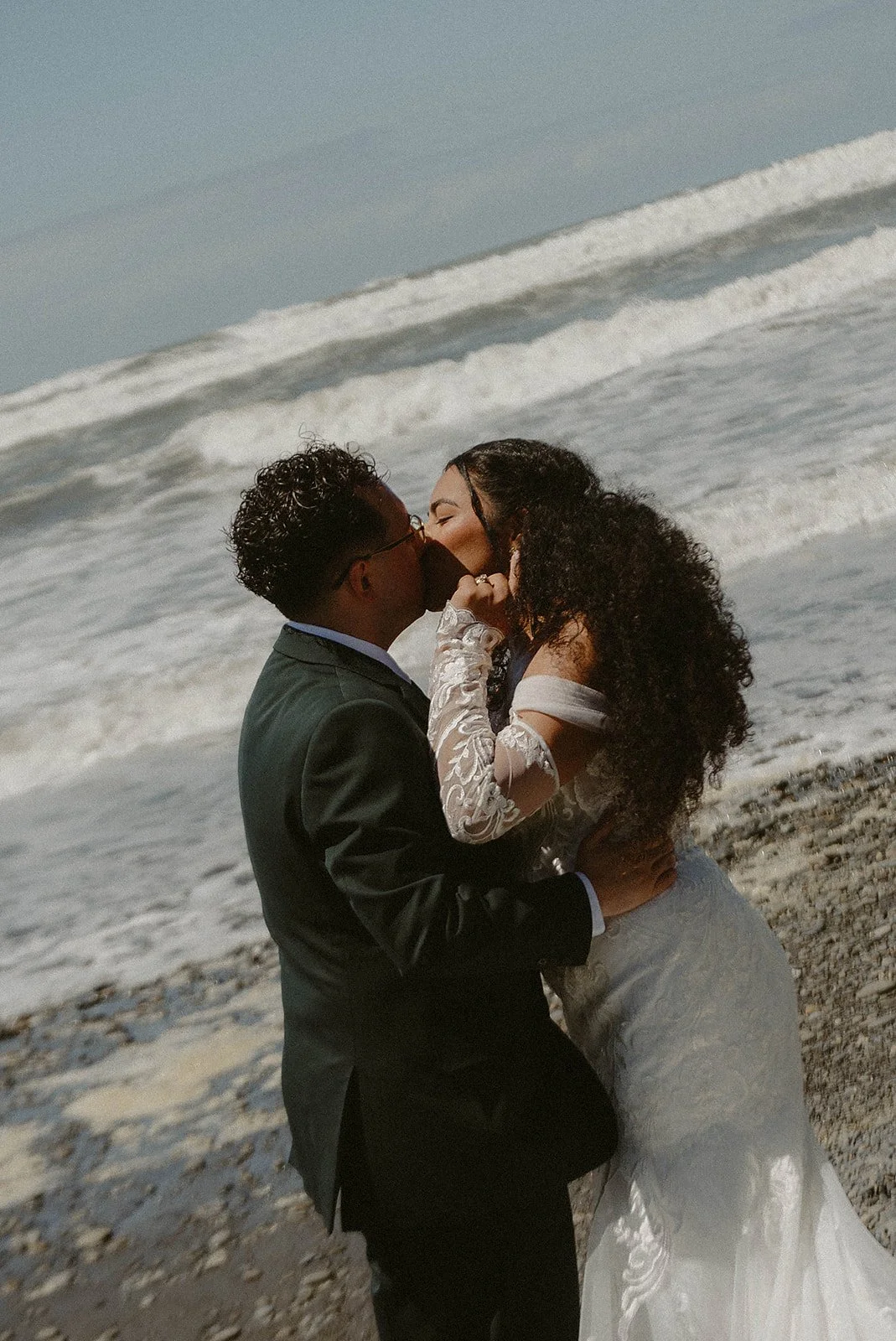 A couple in wedding attire sharing a kiss on the beach with ocean waves in the background.