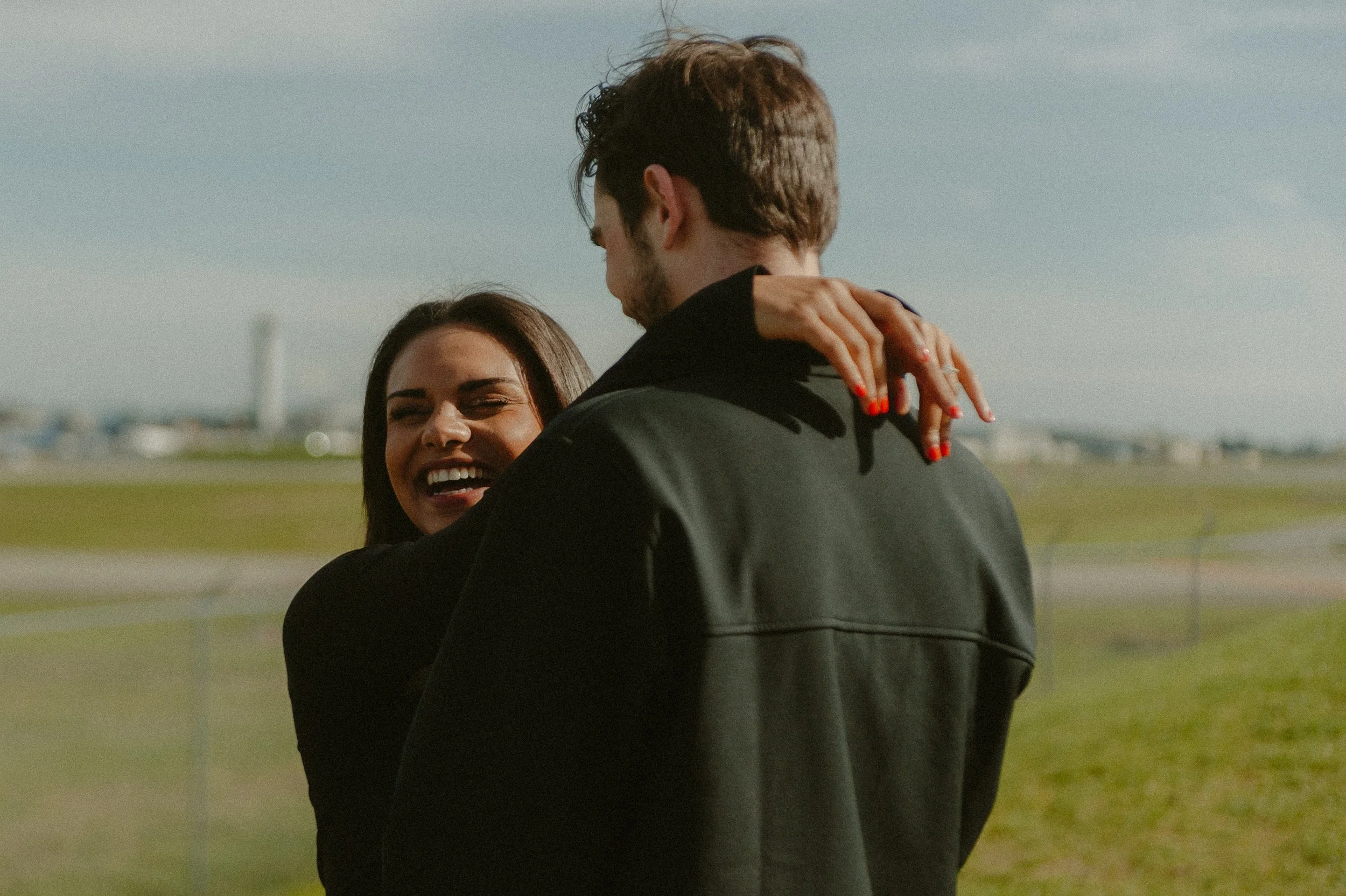 A couple embracing outdoors on a sunny day, with a blurred green field and blue sky in the background.