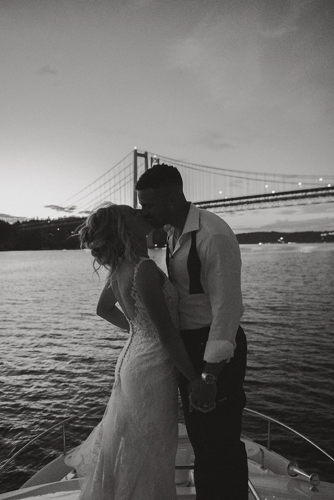 Black and white photo of a couple kissing on a boat at sunset with a bridge in the background.