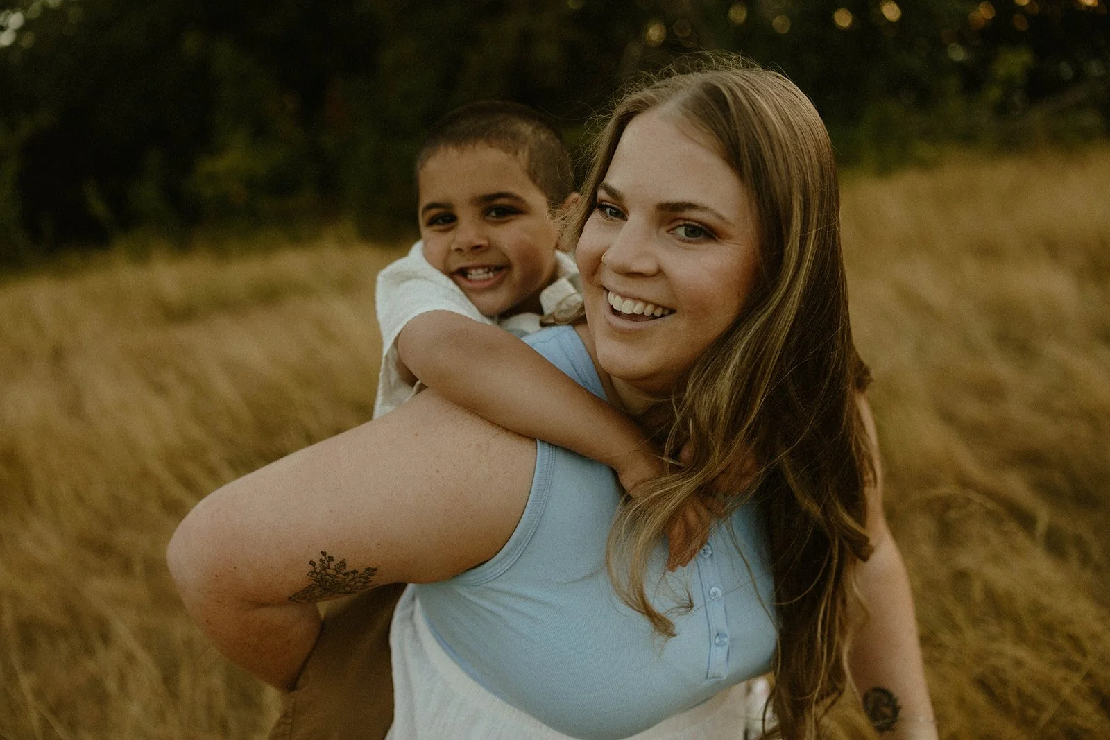 A woman with long brown hair smiling while carrying a young boy on her back in a field of tall grass.