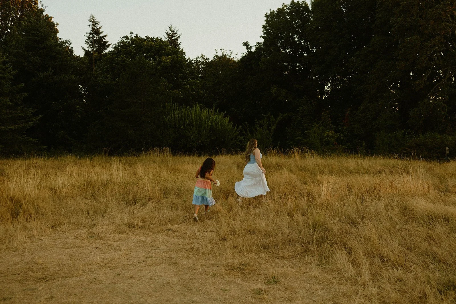 A woman and a young girl walking through a grassy field with trees in the background.