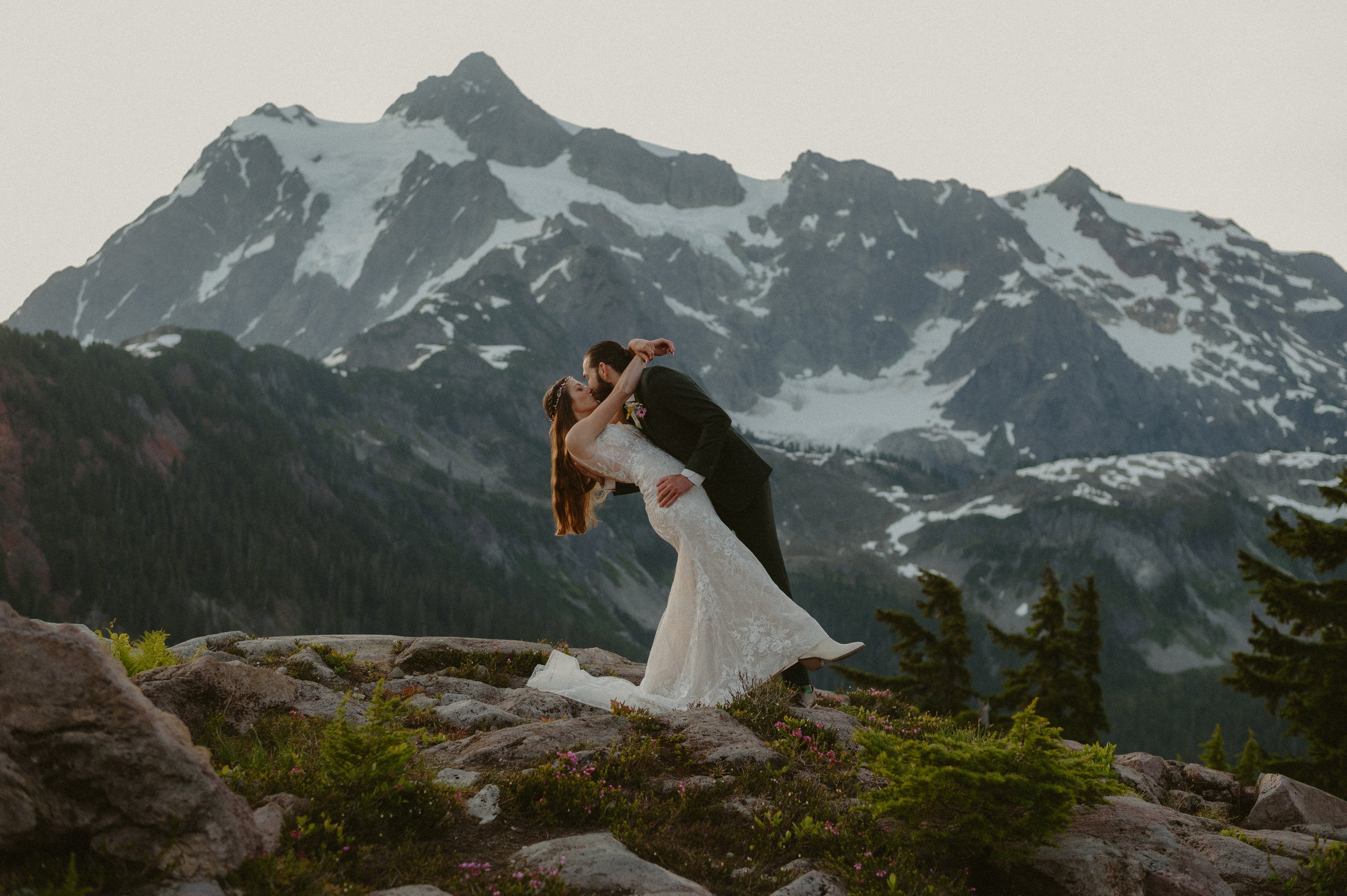 A bride and groom dancing on rocky terrain with snow-capped mountains and pine trees in the background.