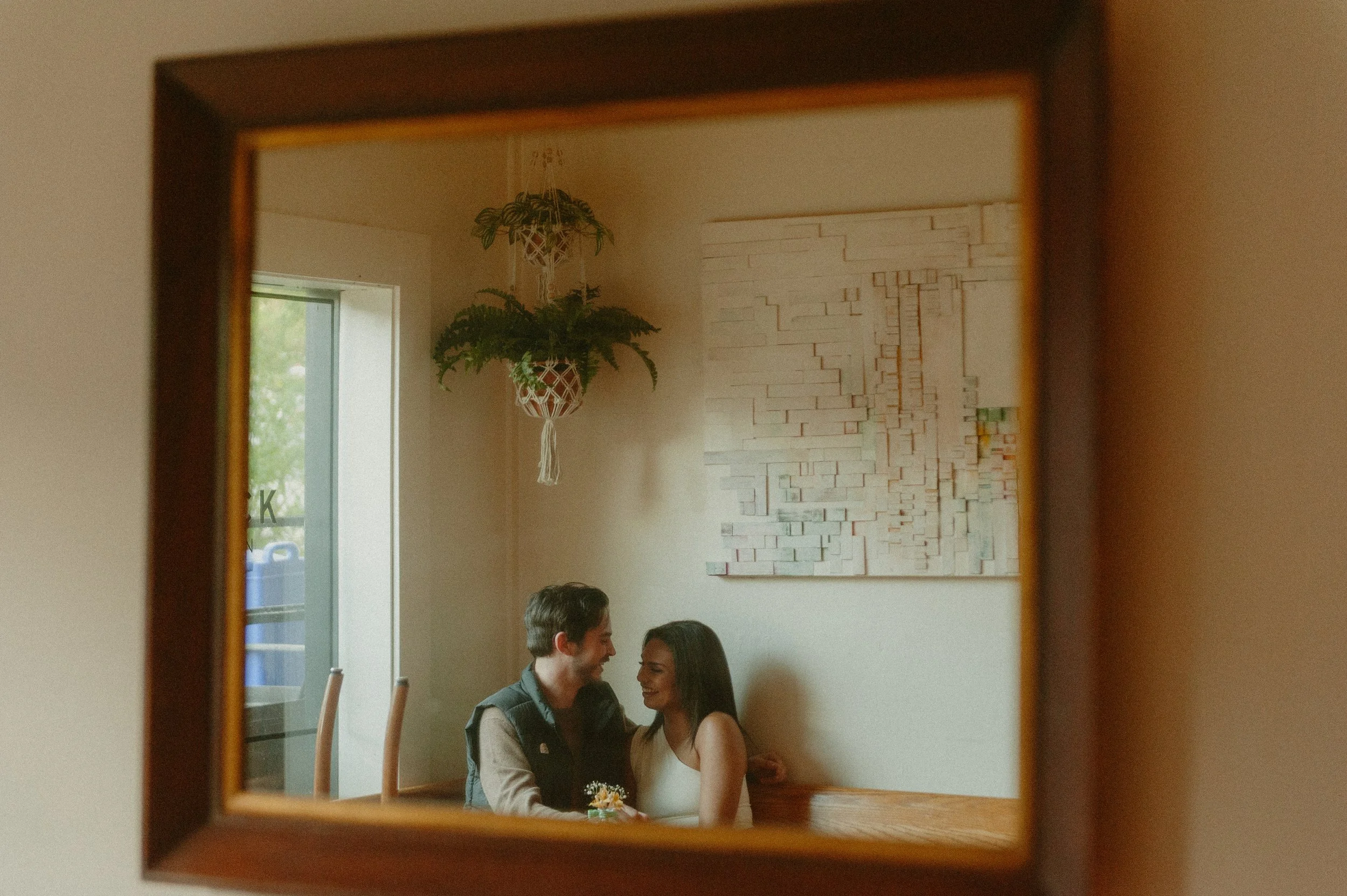 A reflection in a mirror shows a couple sitting close together, smiling at each other. The room has a hanging plant, a window with sunlight coming in, and an abstract textured artwork on the wall.