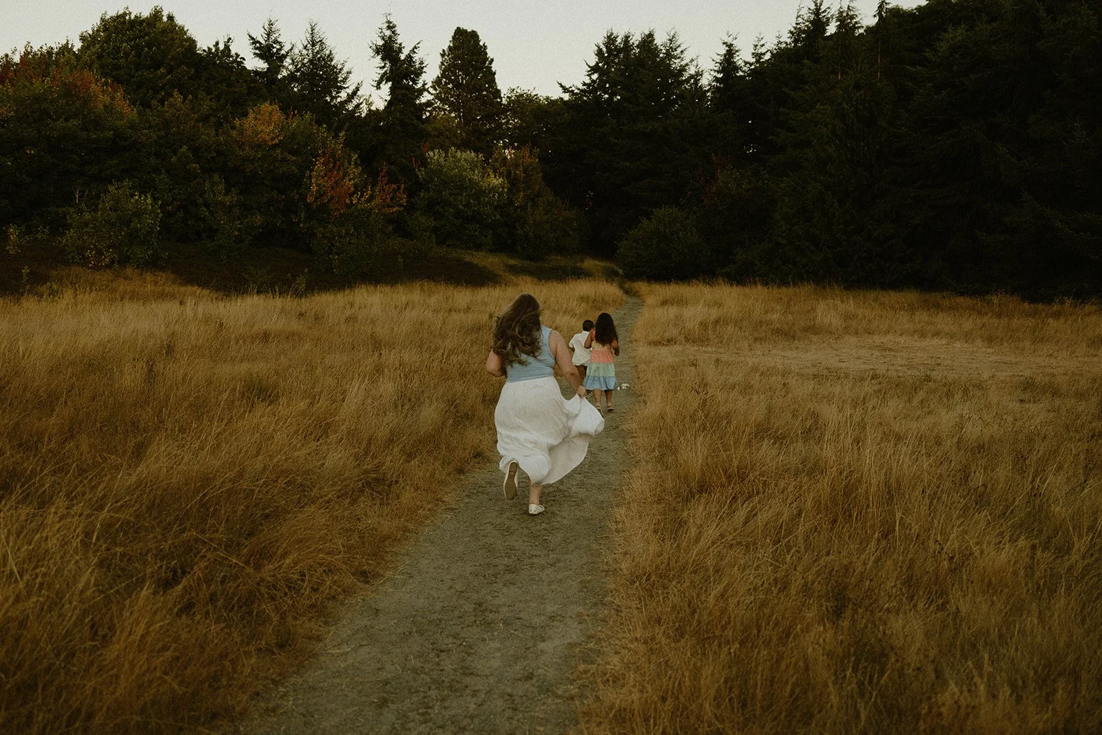 A woman and three children running down a dirt trail through a grassy field surrounded by trees.