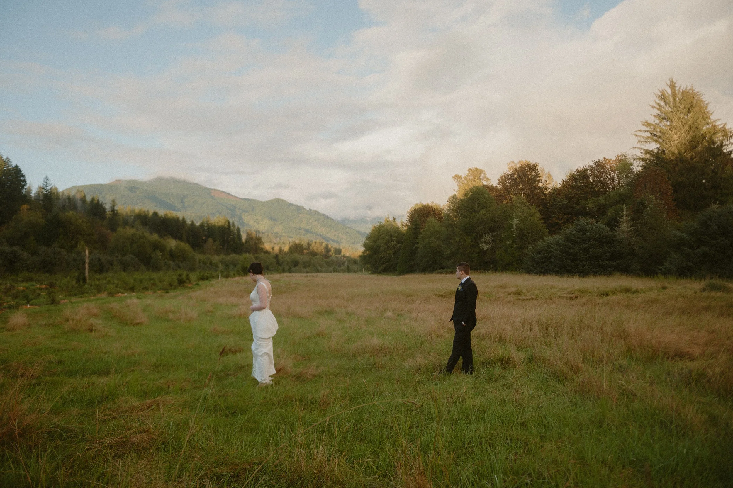 A bride and groom standing apart in a grassy field with trees and mountains in the background during sunset.