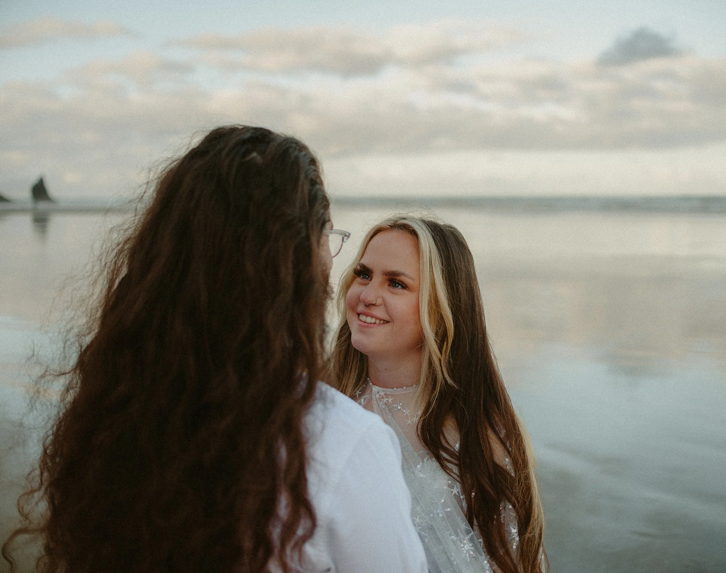 Two women standing on a beach, one with curly dark hair and glasses, the other with long light brown hair, smiling at each other under a cloudy sky.