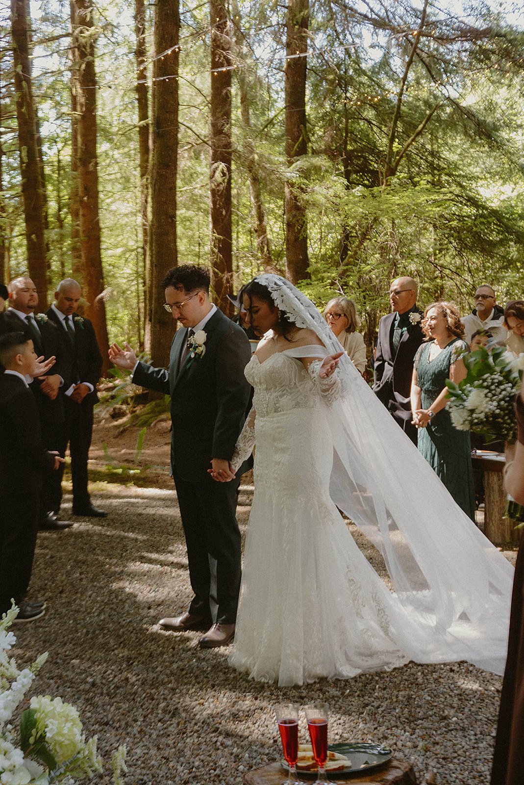 A bride and groom hold hands during their outdoor wedding ceremony in a forest, surrounded by guests.