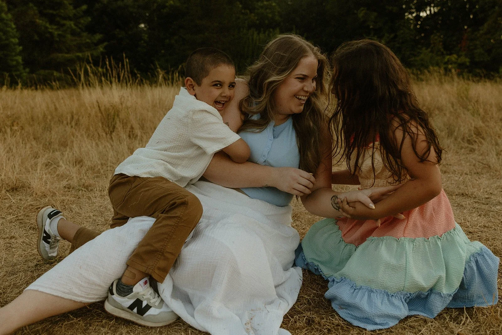A woman with two children, a boy and a girl, playing and laughing together outdoors on a grassy field with trees in the background.