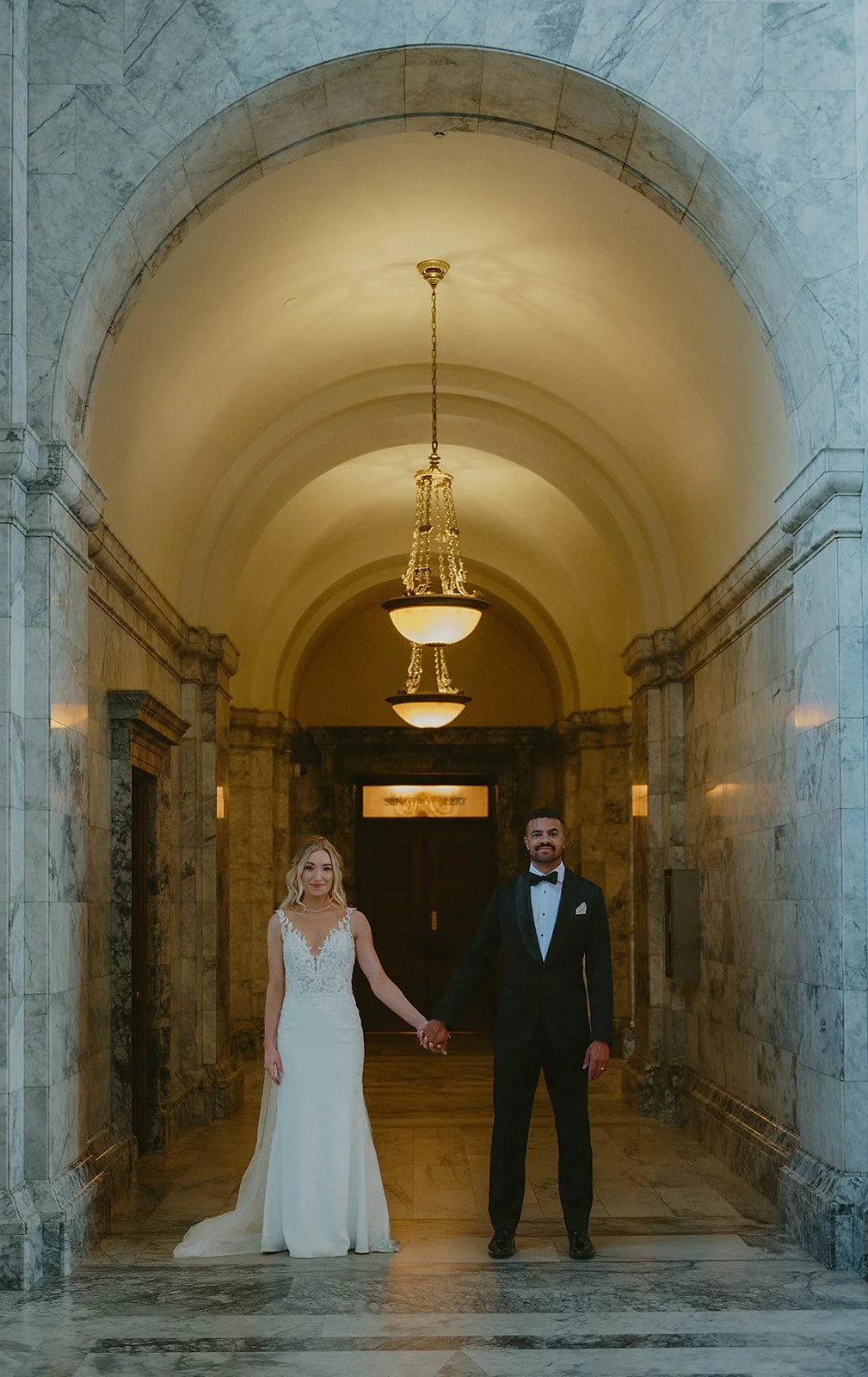 A bride and groom holding hands in a marble hallway with high arched ceiling and pendant lights.