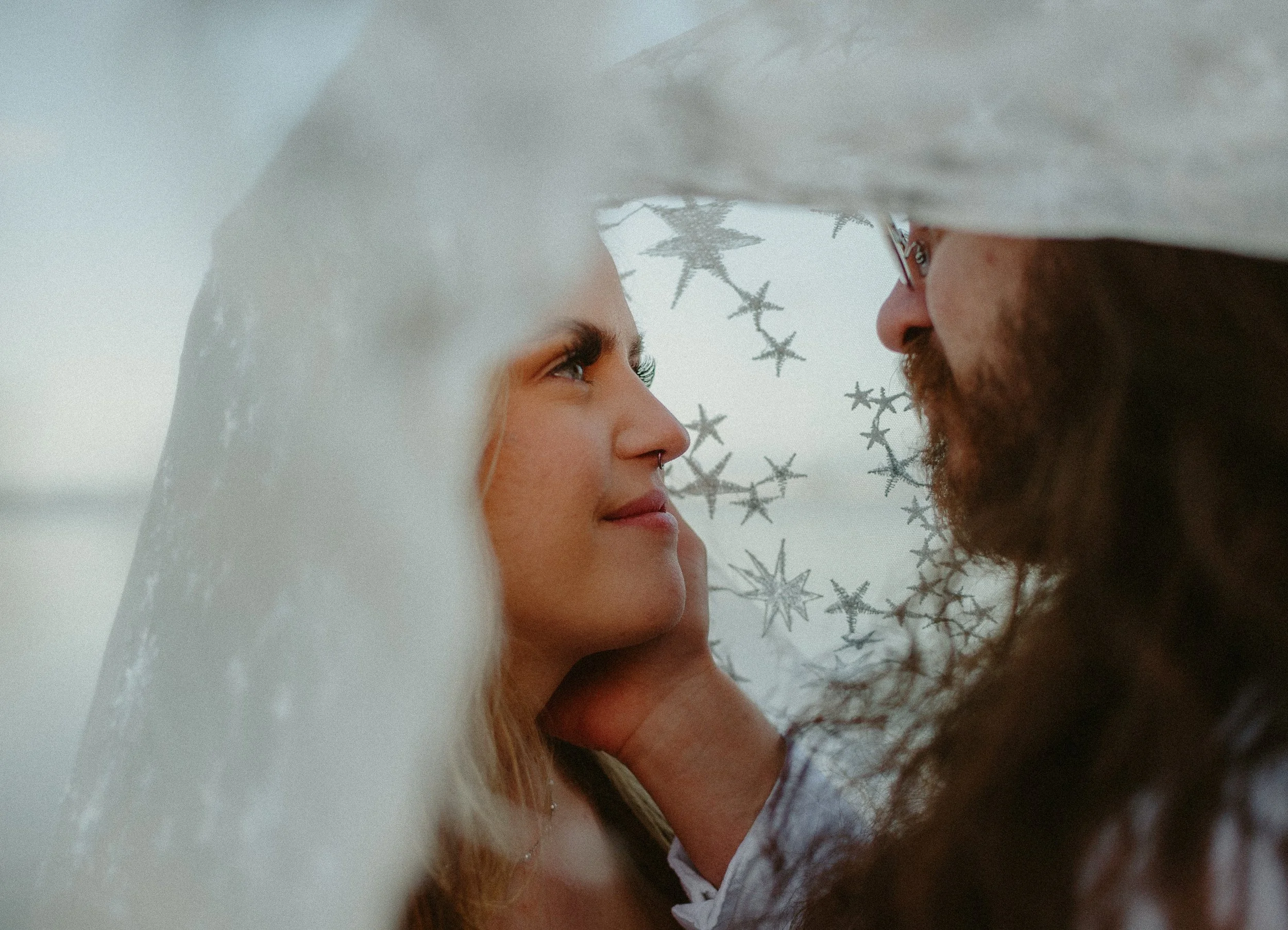 A couple gazing at each other through a curtain of white fabric, with snowflake decorations in the background.