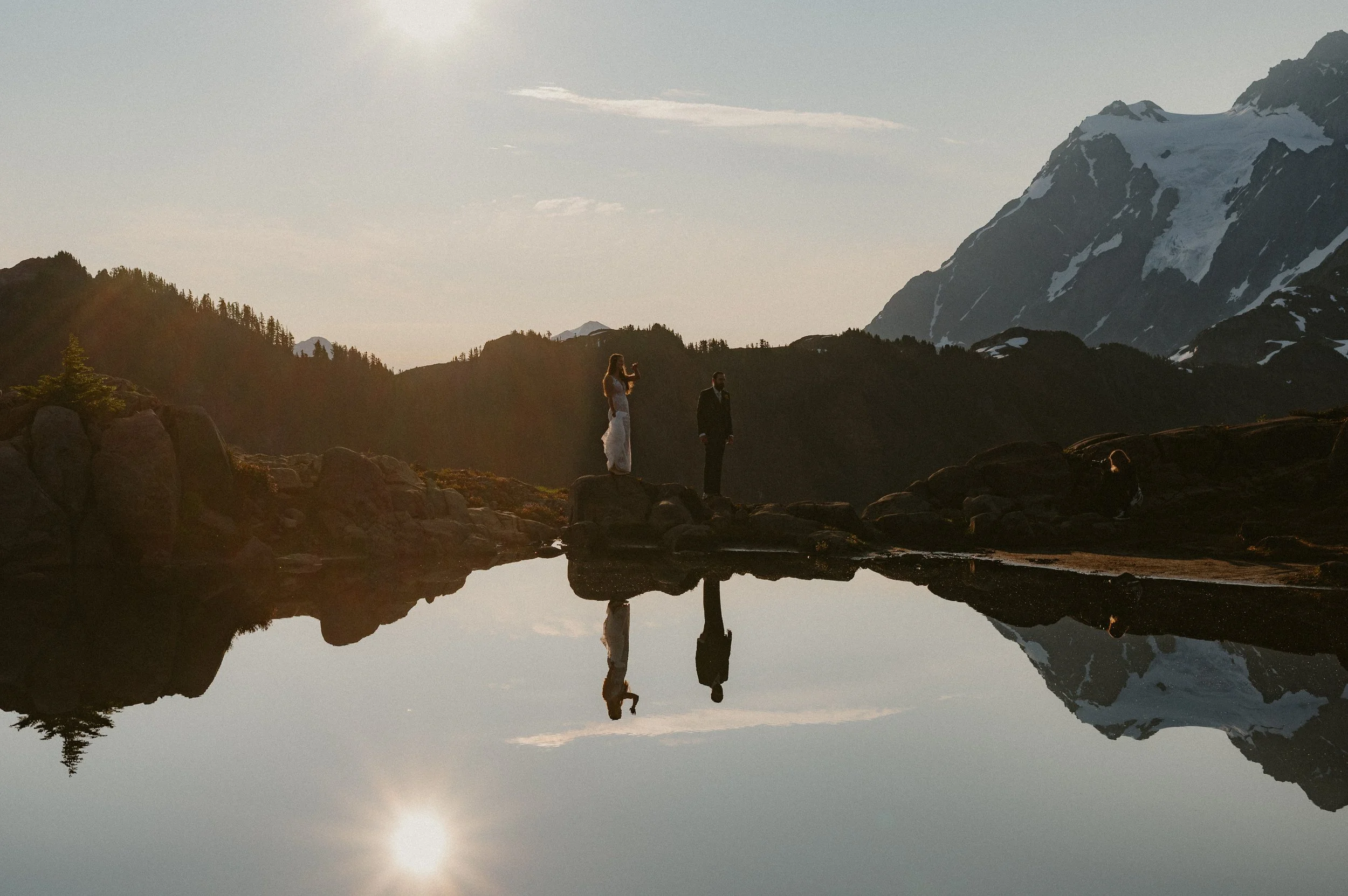 A couple in wedding attire standing on rocks beside a mountain lake with their reflection visible in the water, mountain range in the background, and the sun low in the sky.