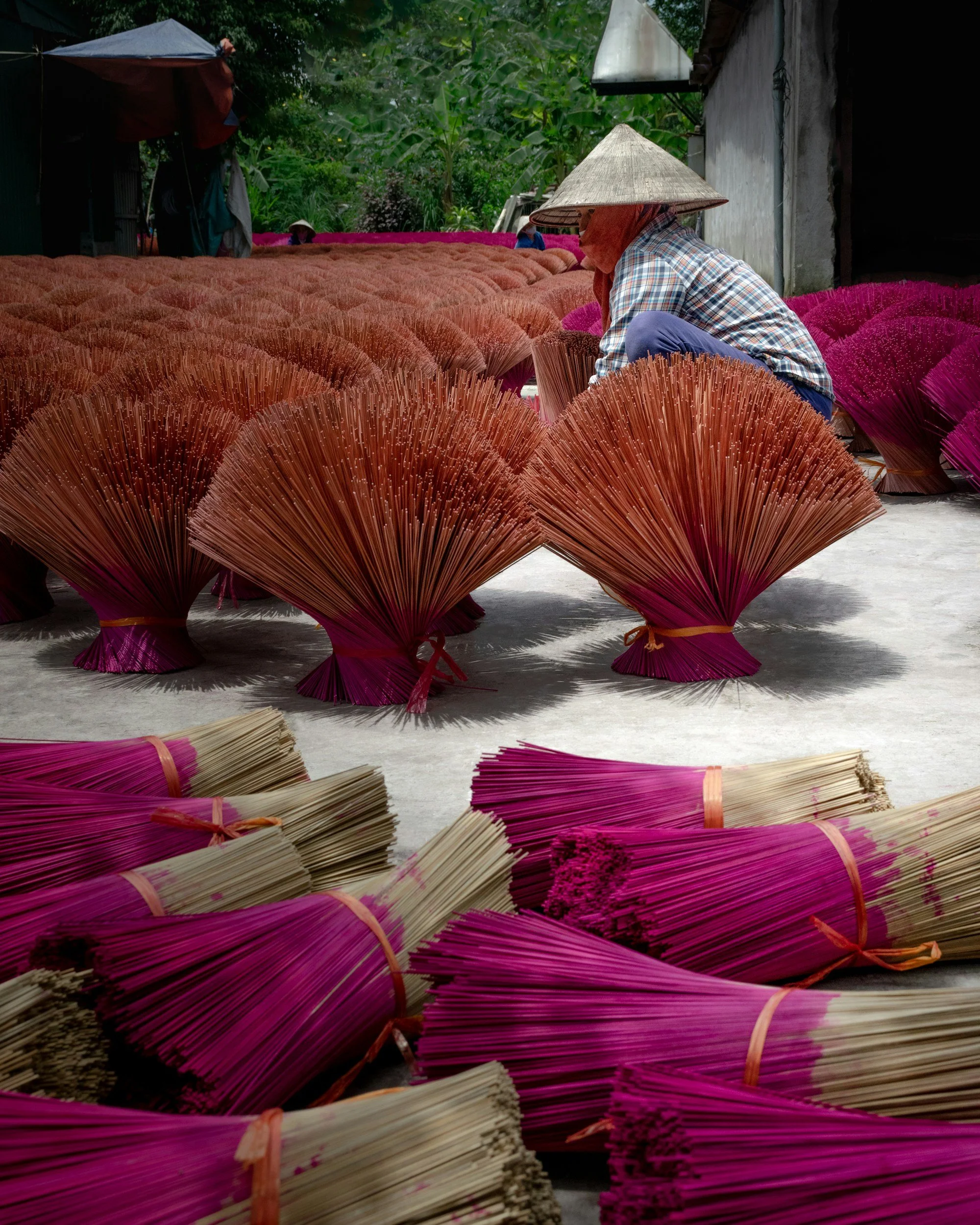 A person wearing a conical hat and a checkered shirt is sitting among bundles of pink and beige straw incense sticks. The individual appears to be sorting or arranging the sticks, with a background of lush green trees and a gray building wall.