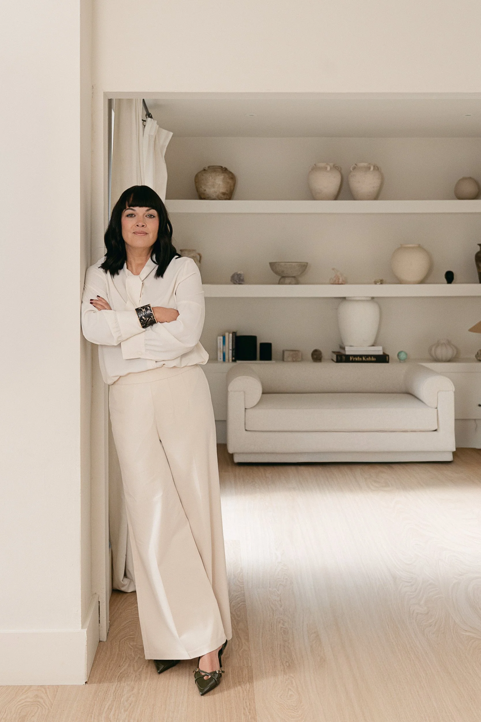A woman standing next to a wall with her arms crossed, wearing a white blouse and light-colored pants, with black and gold shoes, in a room with white shelves displaying decorative objects like vases and books.