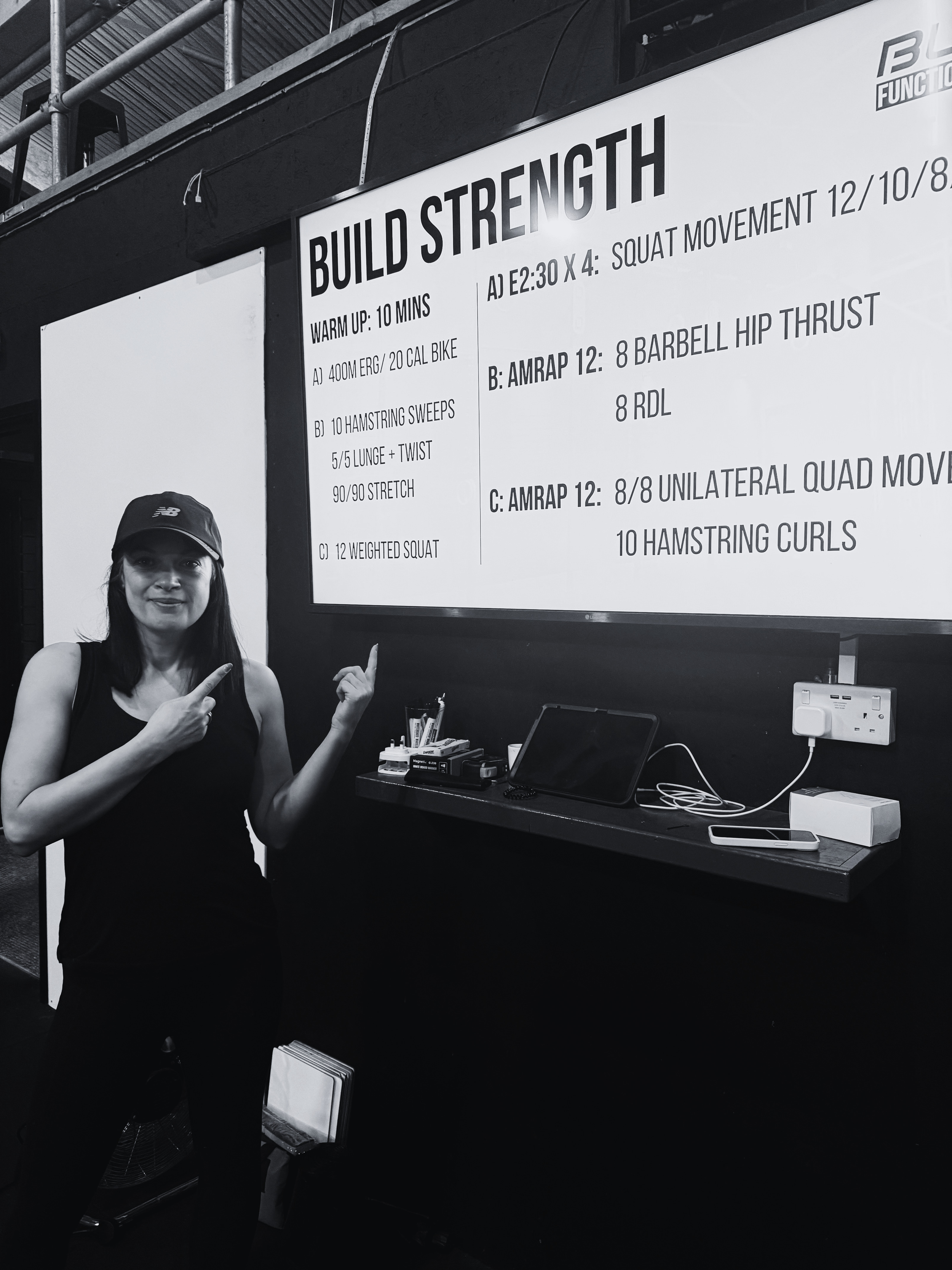 A woman wearing a black cap and sleeveless top is smiling and pointing at a large motivational poster titled 'BUILD STRENGTH' in a gym or fitness area. The poster lists various workout routines and warm-up exercises. There is a desk below the poster with a tablet, some stationery, a smartphone, and electronic devices.