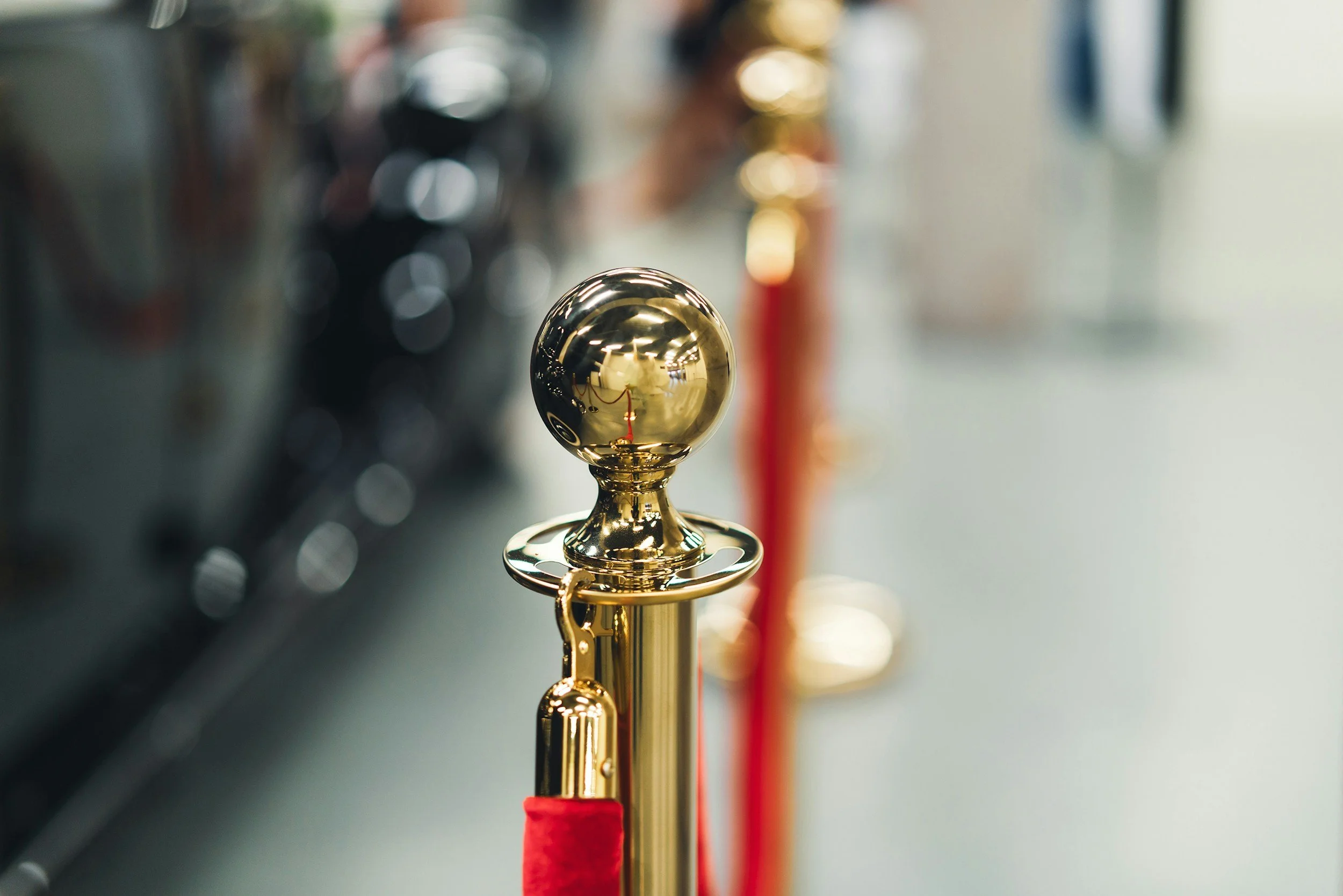 Close-up of a shiny, gold-colored post and ball barrier, with a red velvet rope attached, in an indoor setting.