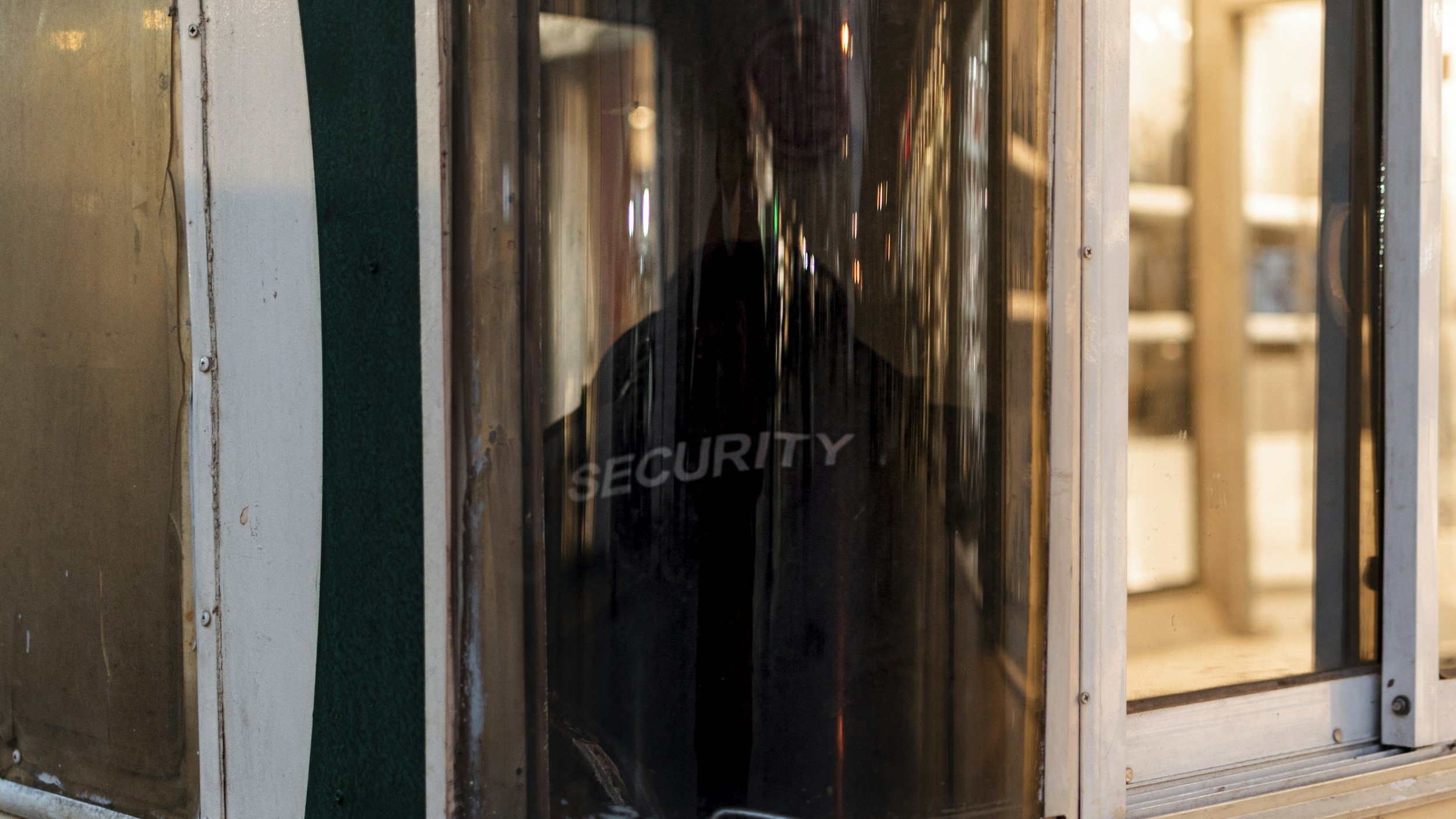 A person wearing a black security jacket standing behind a glass security door inside a building with wooden and metal structures.