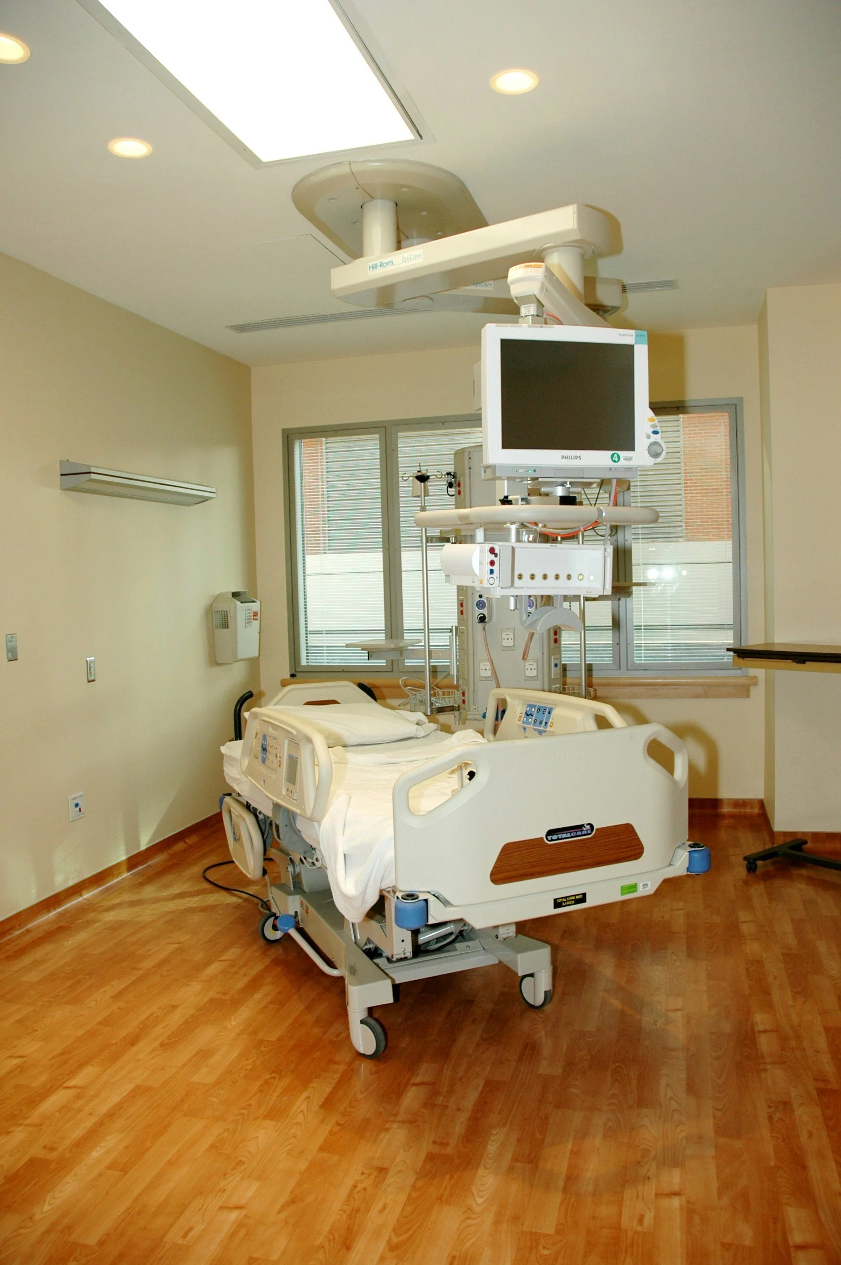 A hospital room with a hospital bed and medical equipment, including a monitor and IV stand, in front of windows with blinds.