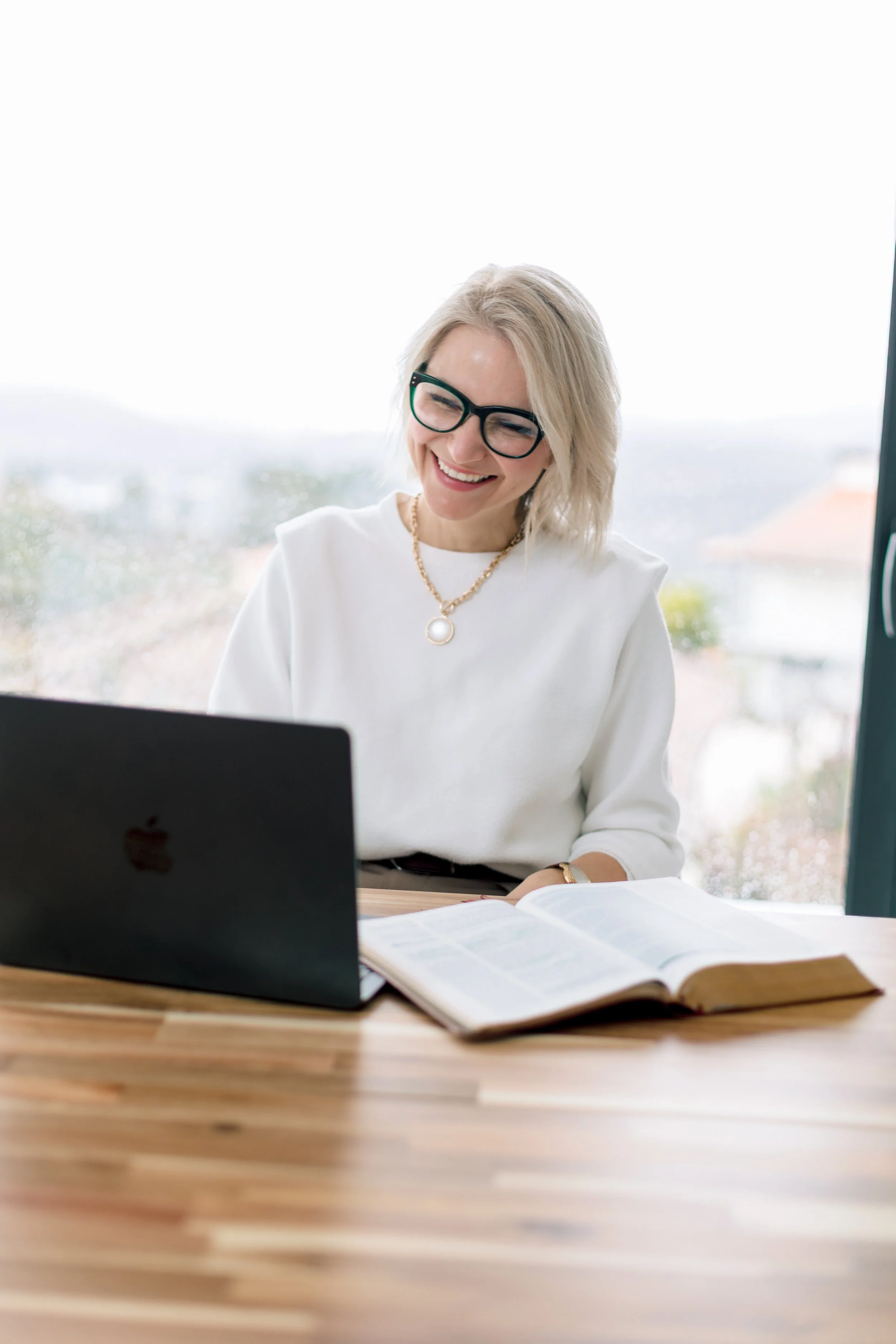 A smiling blonde woman wearing glasses and a white blouse working at a wooden desk with a laptop and open book, sitting by a window with a rainy view outside.