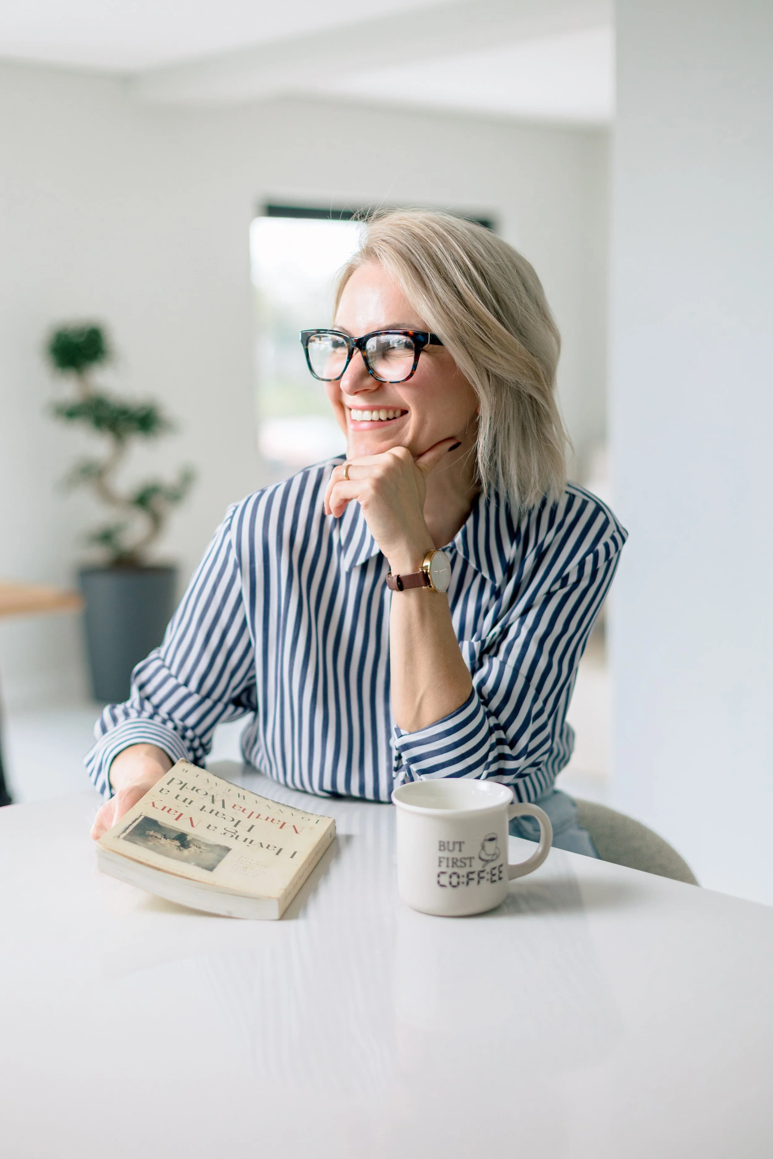 A woman with blonde hair and glasses is smiling and sitting at a table with a book and a coffee mug that says 'But First Coffee'.
