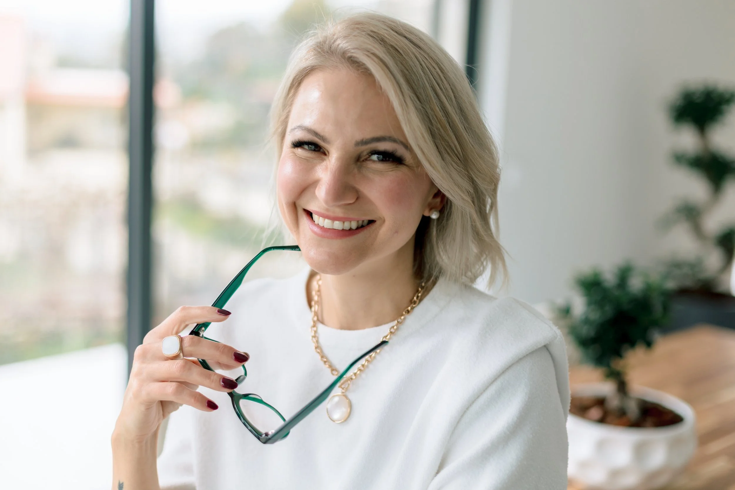 Smiling blonde woman in white shirt holding glasses near her face, indoors with large window and plants in the background.