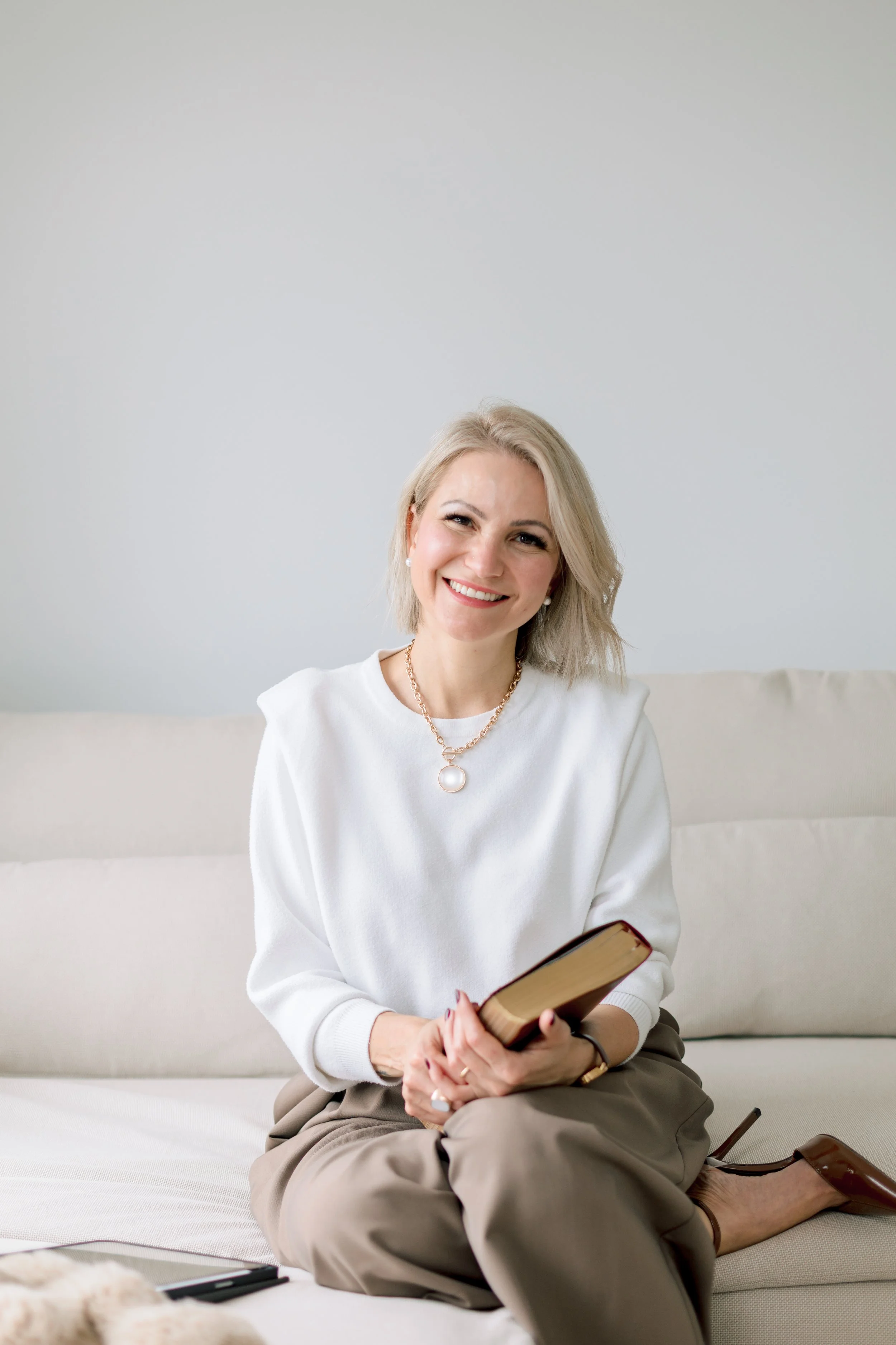 A smiling woman with blonde hair sitting on a beige sofa, holding a closed book, wearing a white sweater, beige pants, and brown heels, in a bright room.