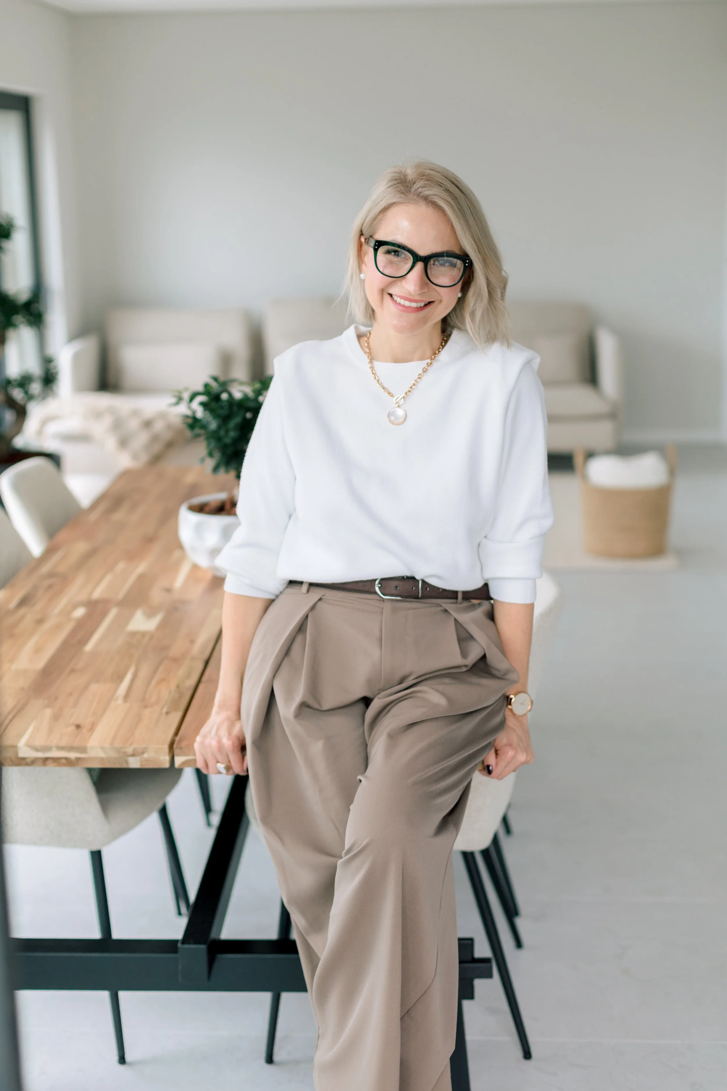 A woman with blonde hair, glasses, and a pearl necklace leaning on a wooden table in a bright, modern dining room.
