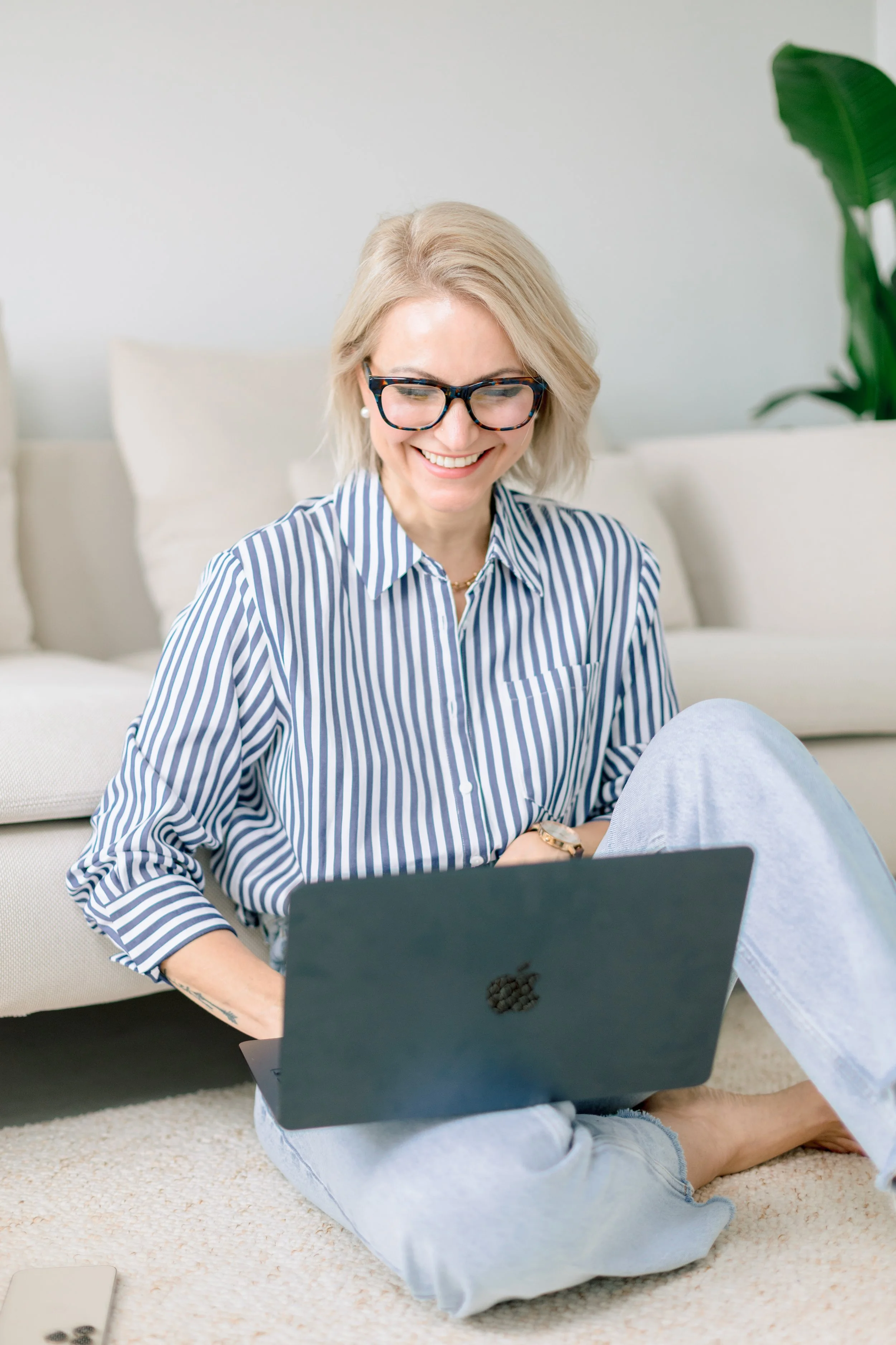 A woman with blonde hair and glasses sitting on the carpet with a laptop, smiling and looking at the screen.