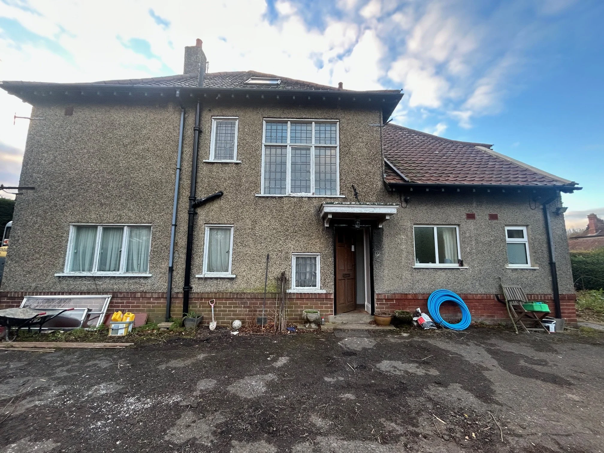 Front view of a two-story house with a rough-textured exterior, large front windows, and a wooden front door. The yard has gardening tools, a garden hose, and some gardening supplies scattered around.