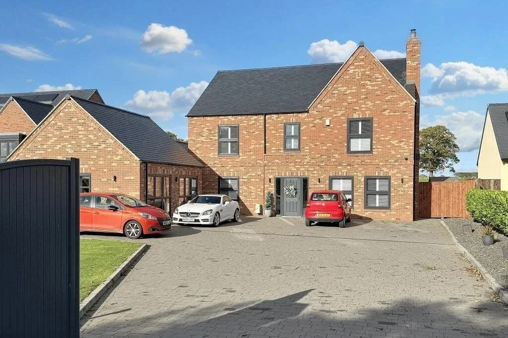 A brick house with a gray front door and four windows, cars parked in the driveway, a blue sky with some clouds, and neighboring houses on each side.