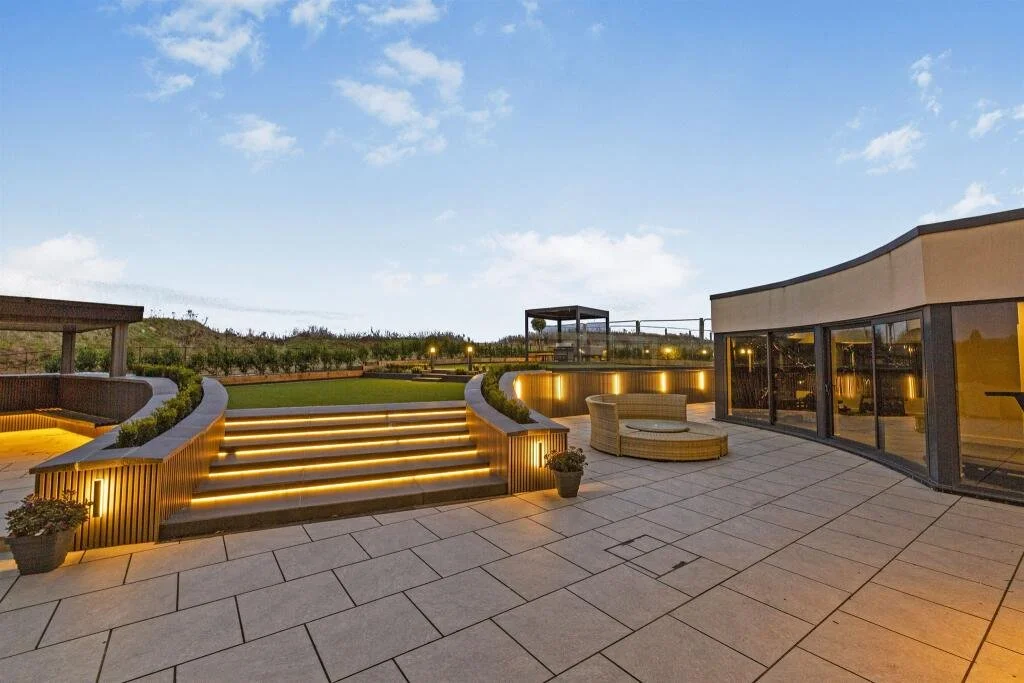 City rooftop terrace with modern outdoor furniture, illuminated stairs, potted plants, and a view of the overlooking cityscape at dusk.