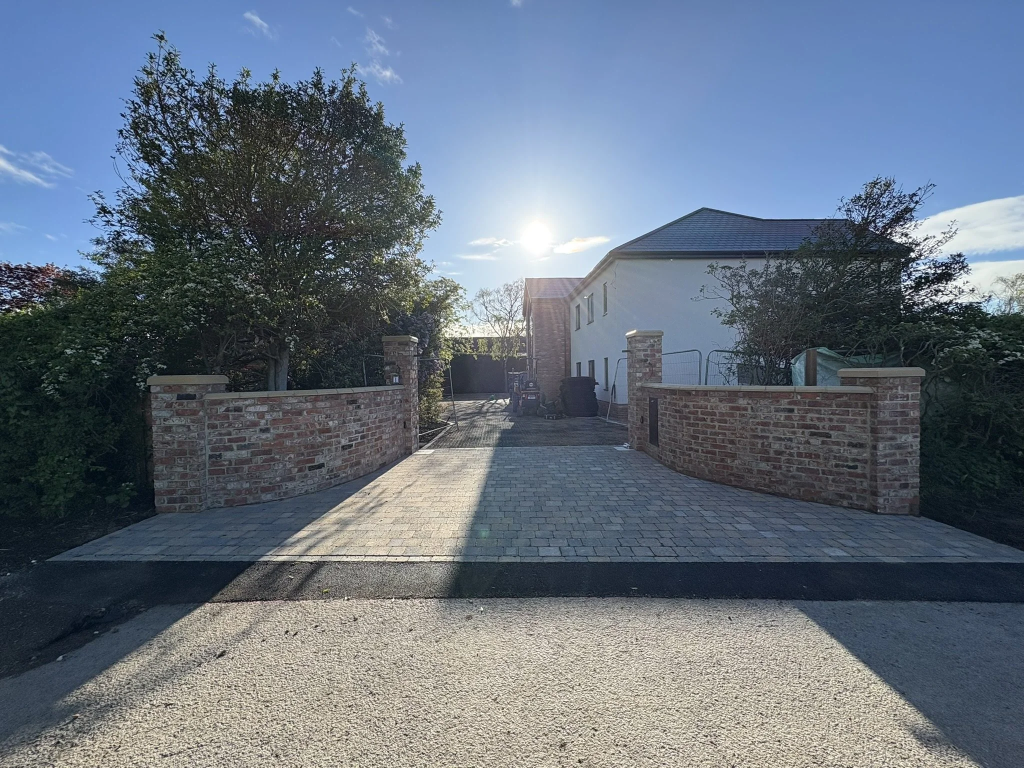 A paved driveway in front of a house with a white exterior and a sloped roof, flanked by brick walls and tall trees on either side, under a clear blue sky with the sun shining.