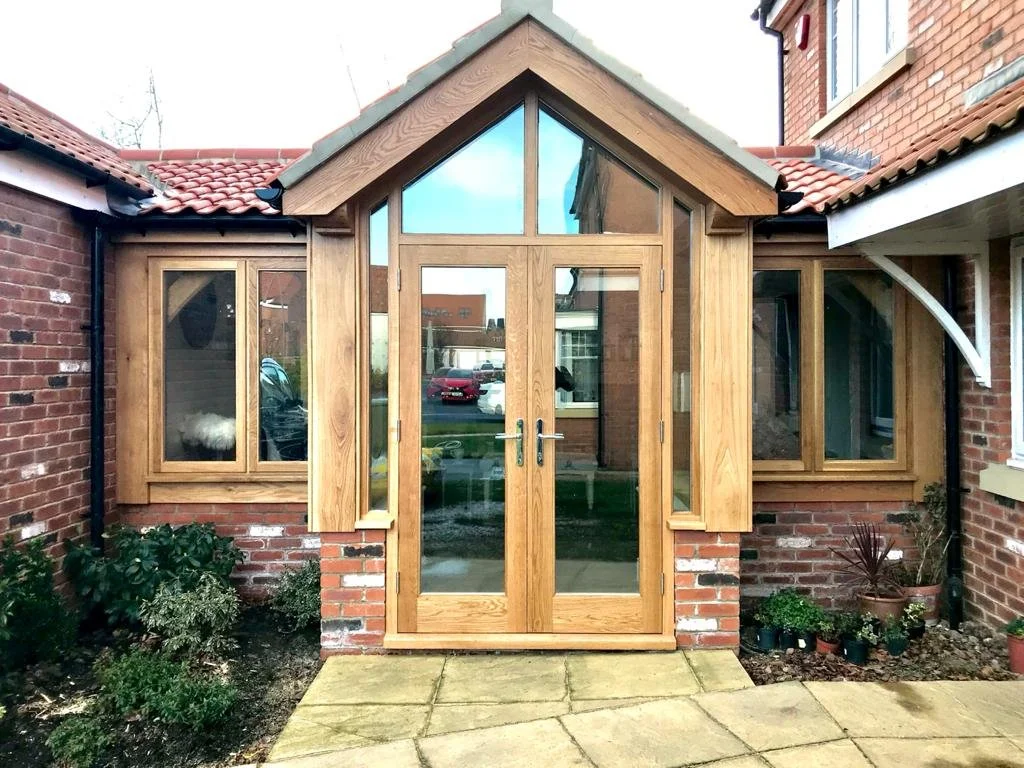 Newly constructed wooden porch and extension with glass doors and windows attached to a brick house with a red tiled roof, surrounded by small shrubs and potted plants.