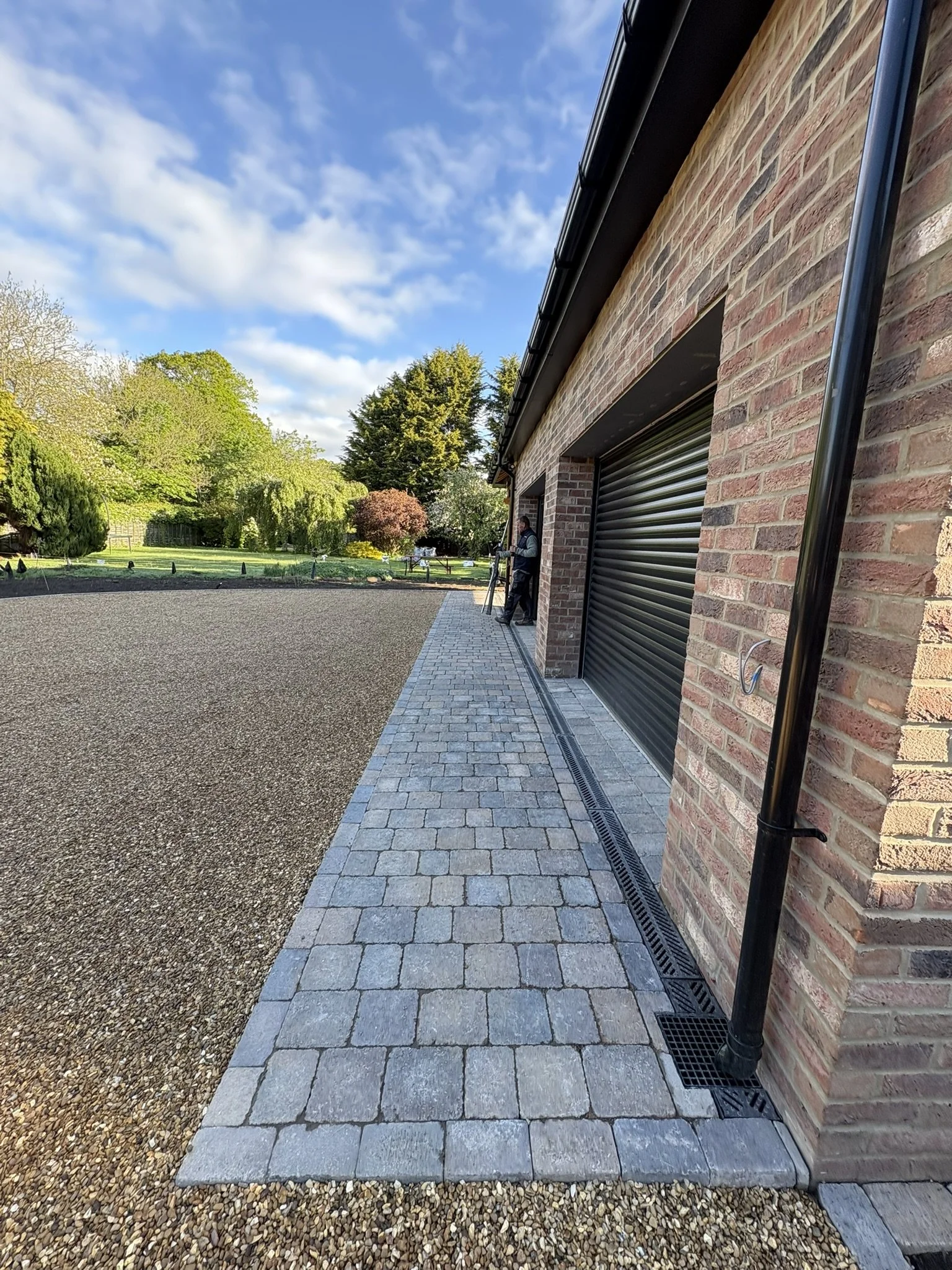 A patio area next to a brick house with a roller door, paved with cobblestones and surrounded by a garden with green trees and shrubs under a blue sky with some clouds.