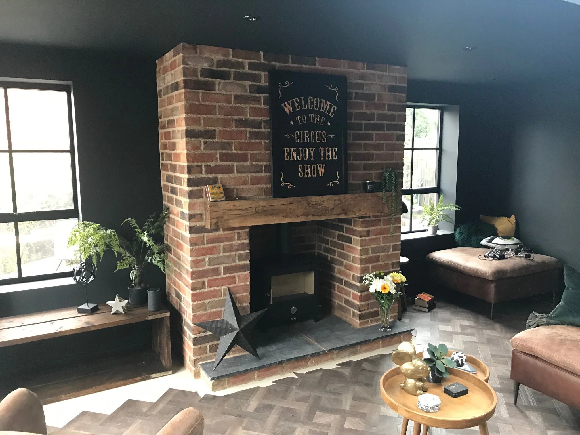 Living room with brick fireplace, black wall, and wooden furniture. Decor includes plants, flowers, and decorative items, with natural light coming through window.
