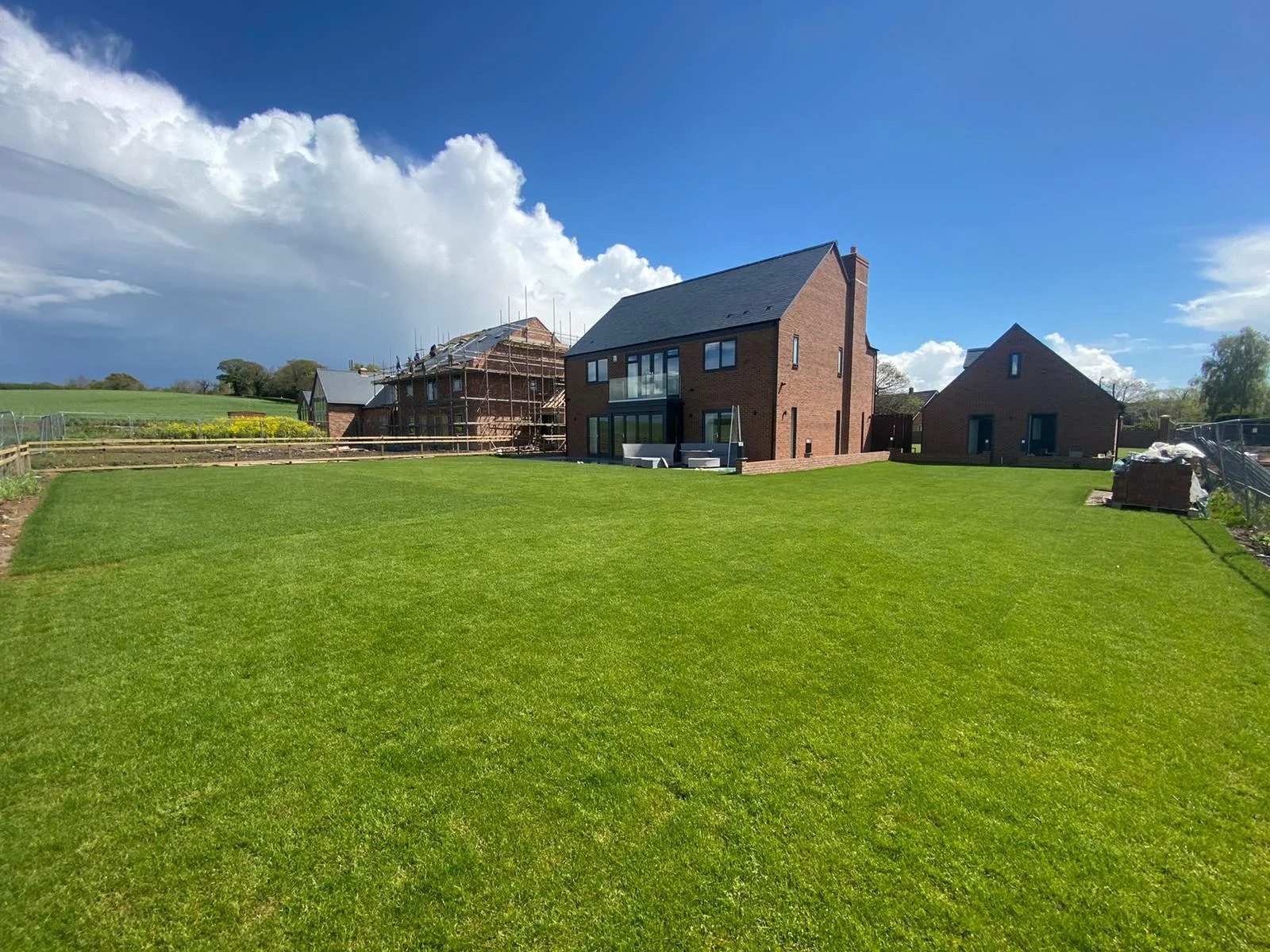 Newly renovated house with brick exterior, large lawn, and construction in progress on neighboring homes under a bright blue sky with some clouds.